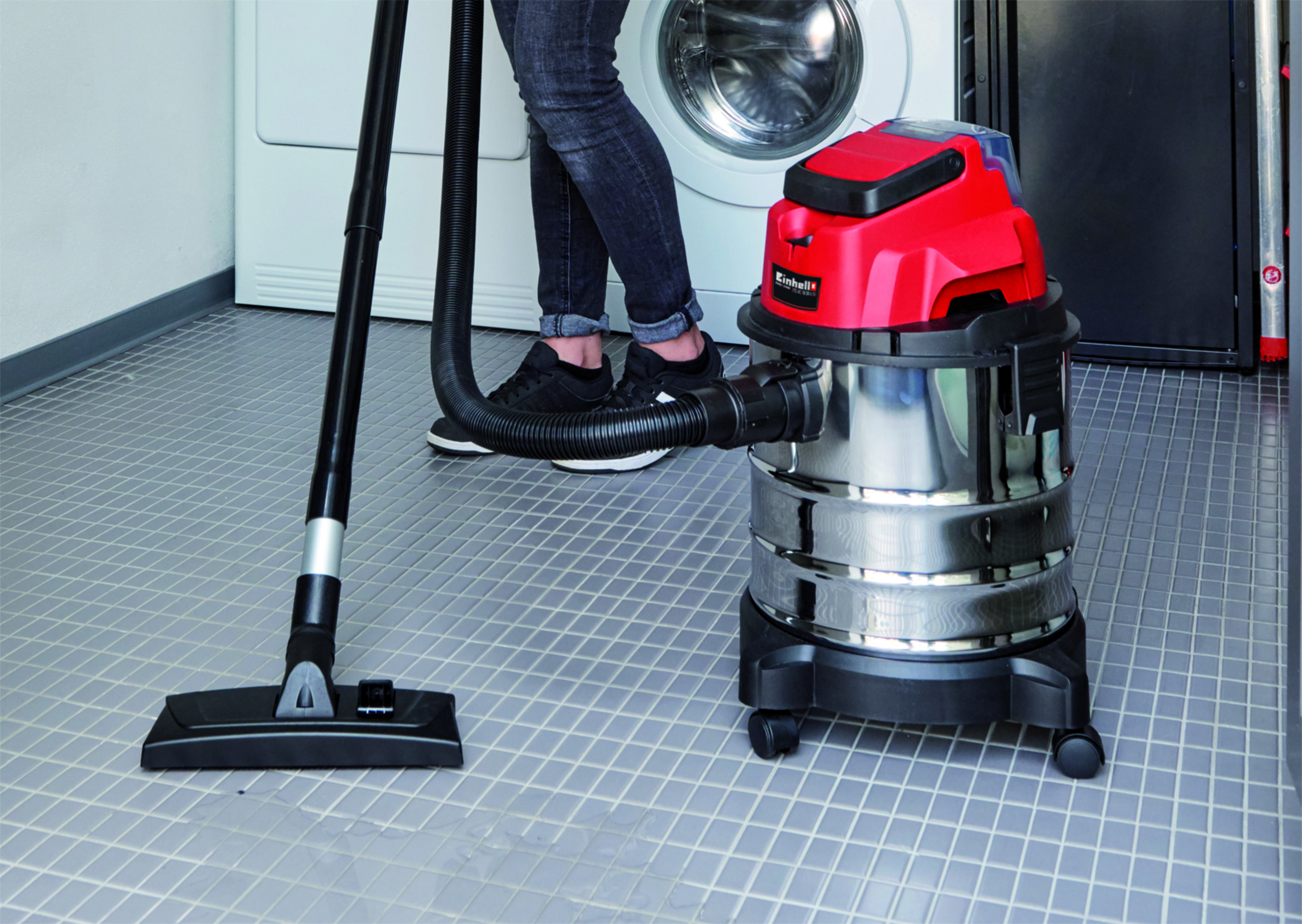 Une personne utilise un aspirateur pour nettoyer un sol carrelé. Un lave-linge est à l'arrière-plan.