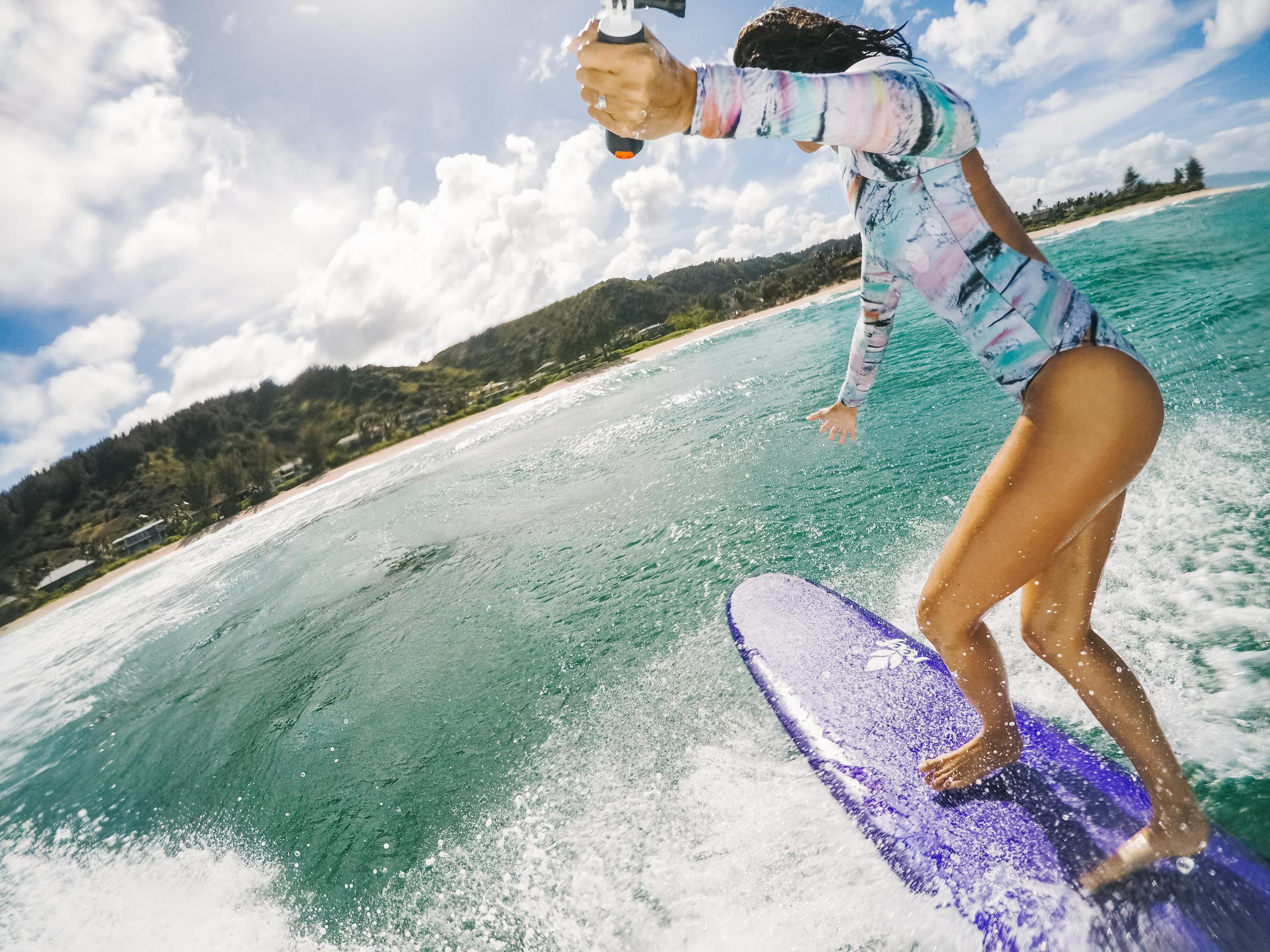 Kobieta surfuje na niebieskiej desce w oceanie, z zachmurzonym niebem i plażą w tle.