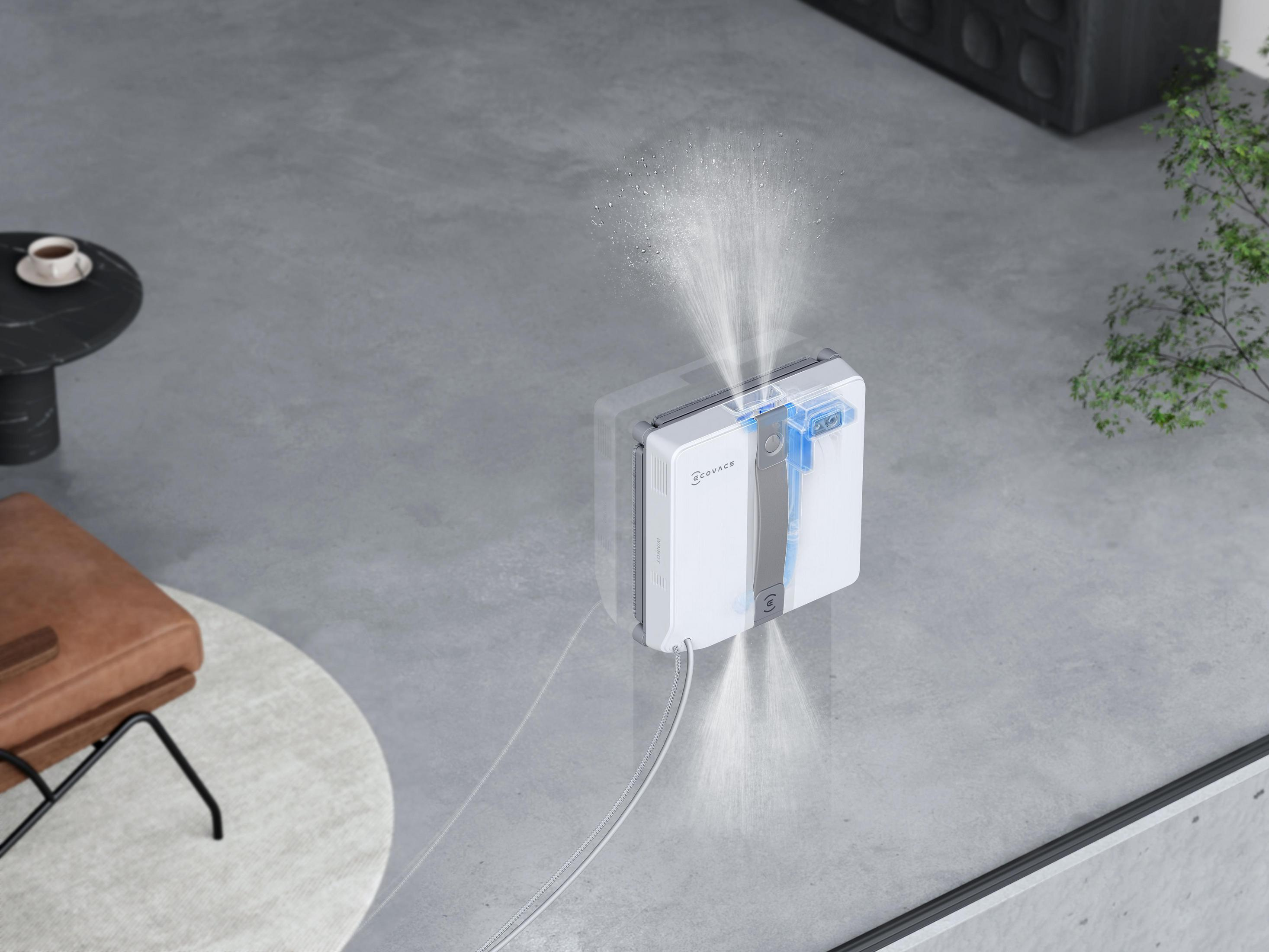 Un robot per la pulizia dei vetri bianco spruzza acqua mentre pulisce una finestra. A sinistra c'è una tazza di caffè.