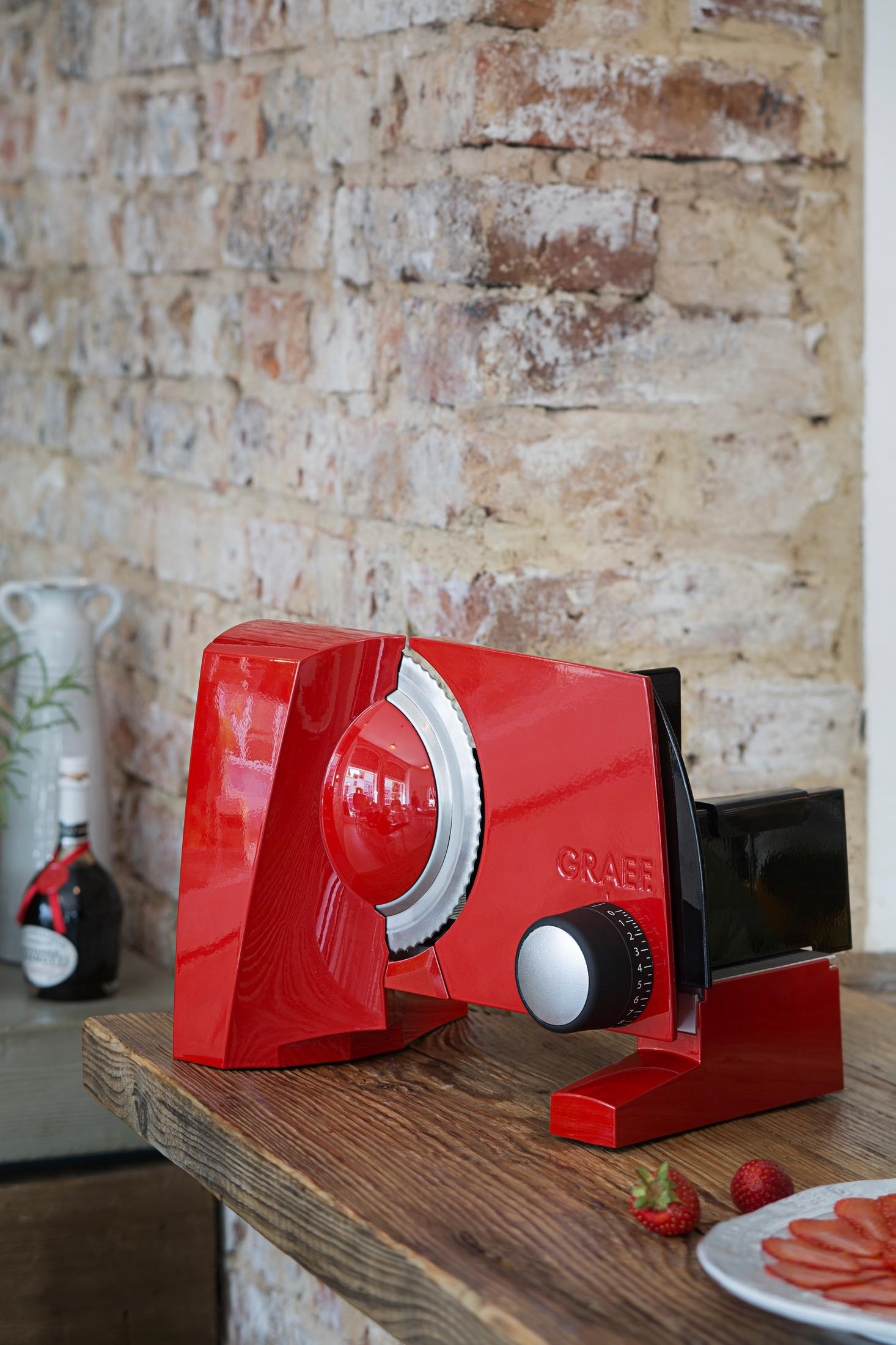 Czerwona krajalnica do żywności na drewnianym stole, truskawki i ceglana ściana w tle.