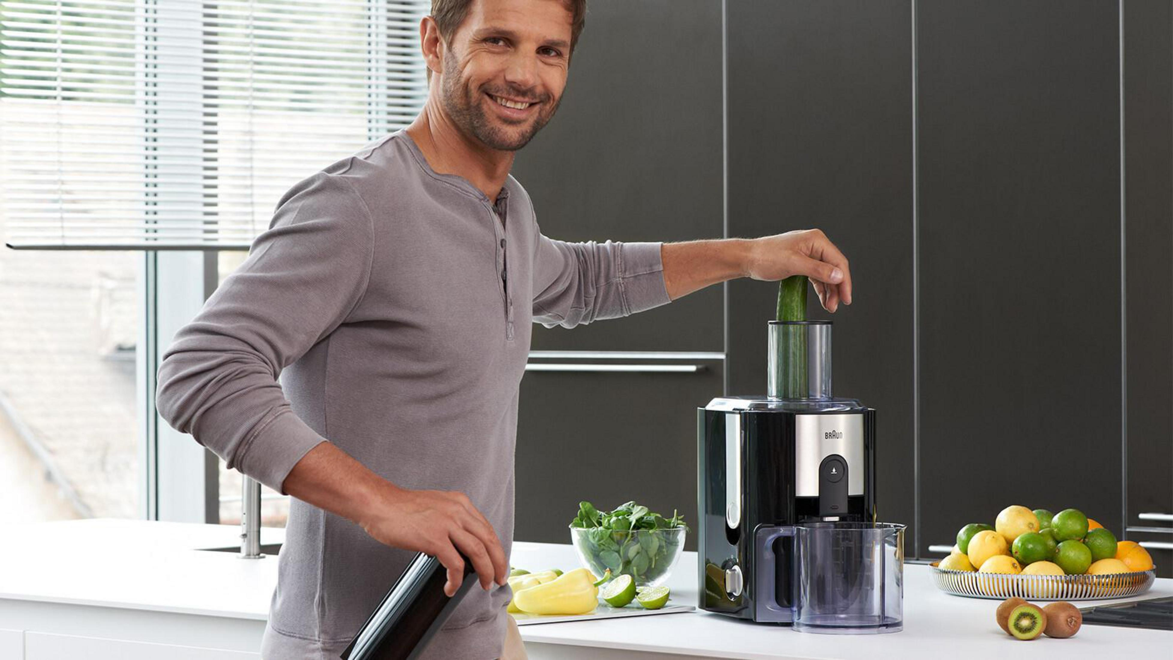 Un hombre está exprimiendo un pepino. Está cerca de limones, kiwi y un exprimidor Braun. Hay un recipiente negro.