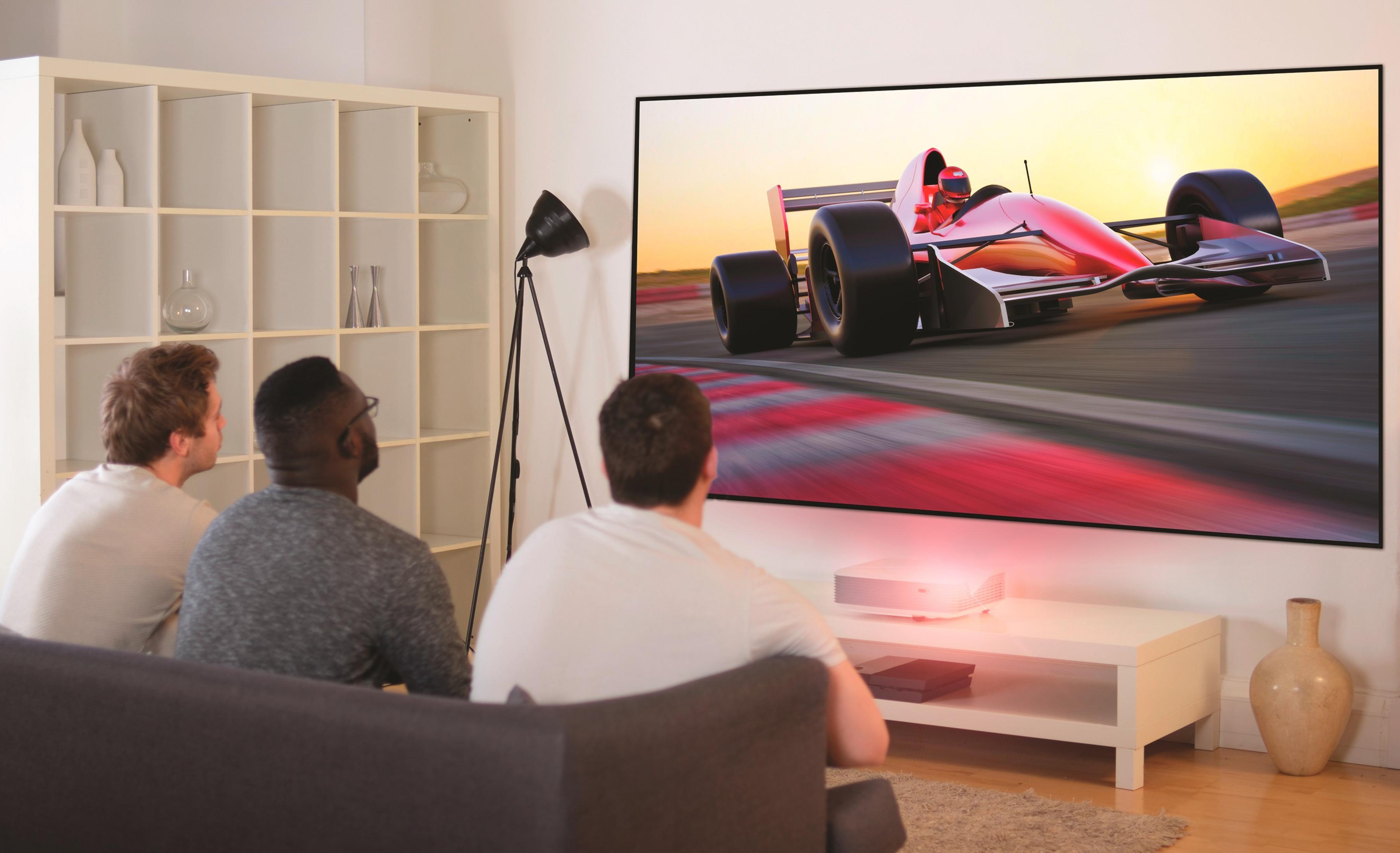 Tres hombres viendo un coche de carreras en una pantalla grande. Un mueble blanco al frente.