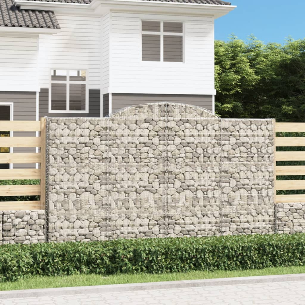 Gabionen-Steinmauer vor einem Haus. Ein Holzzaun und Büsche sind ebenfalls sichtbar.