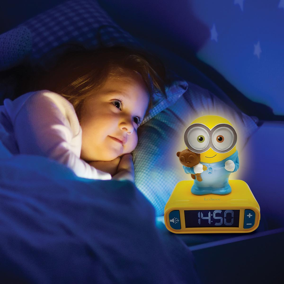 Una niña mira una luz nocturna con forma de Minion. El despertador muestra 14:50. La habitación está oscura.