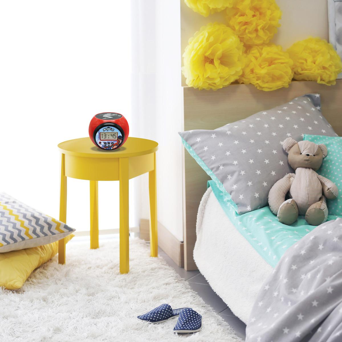 Un despertador Miraculous sobre una mesa amarilla en un dormitorio. Se ve una cama con almohadas y un oso de peluche.