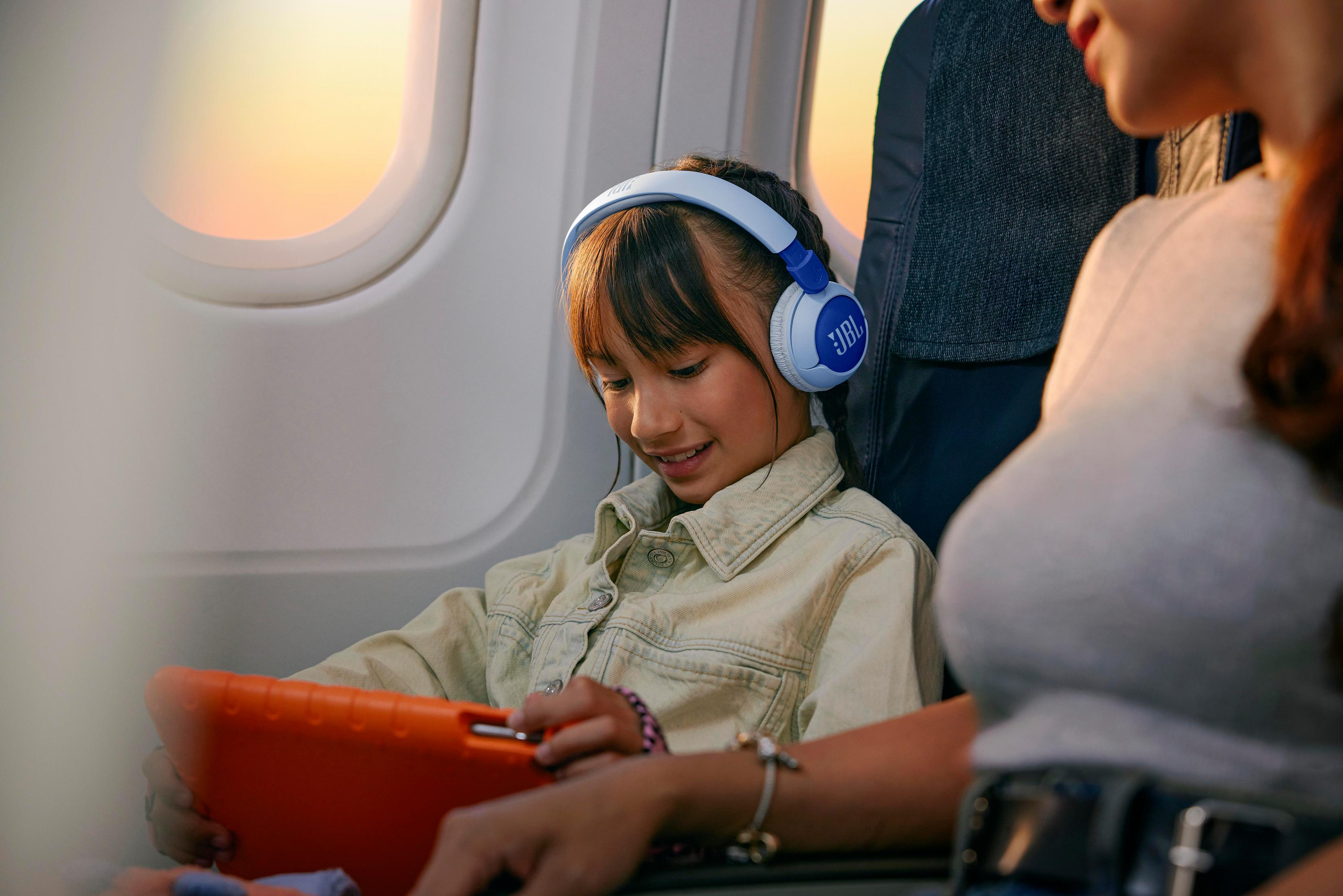 Ragazza in aereo con cuffie e tablet. Donna accanto a lei. Cielo luminoso fuori.