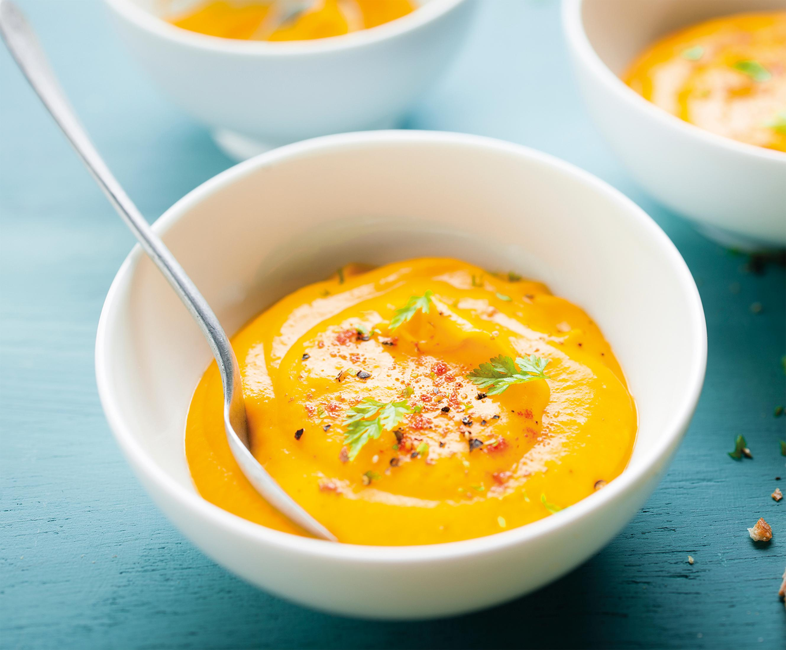 Biała miska z pomarańczową zupą, udekorowana ziołami, na niebieskiej powierzchni.