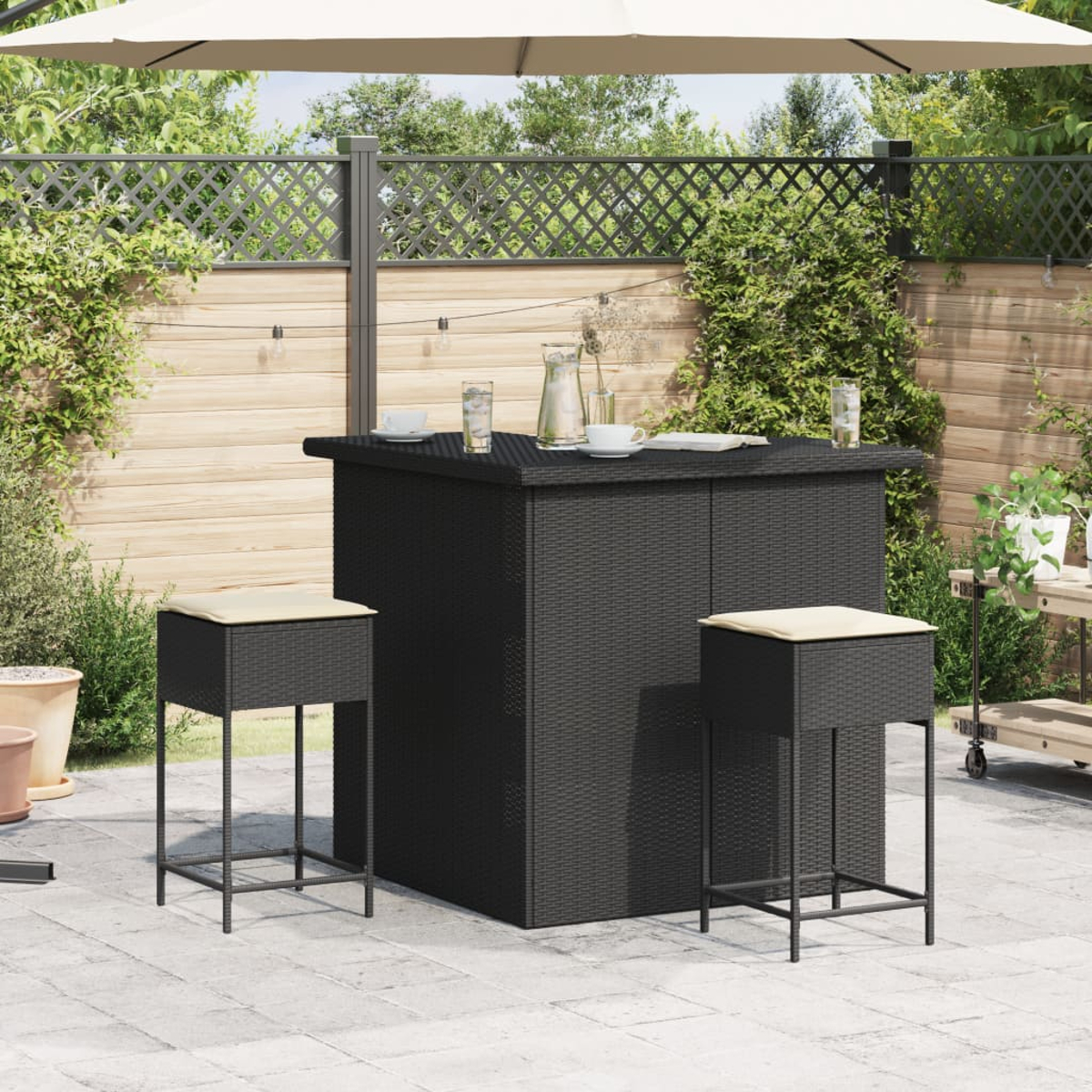 Schwarze Rattan-Bar mit Hockern. Sonnenschirm im Hintergrund, mit Pflanzen drumherum.