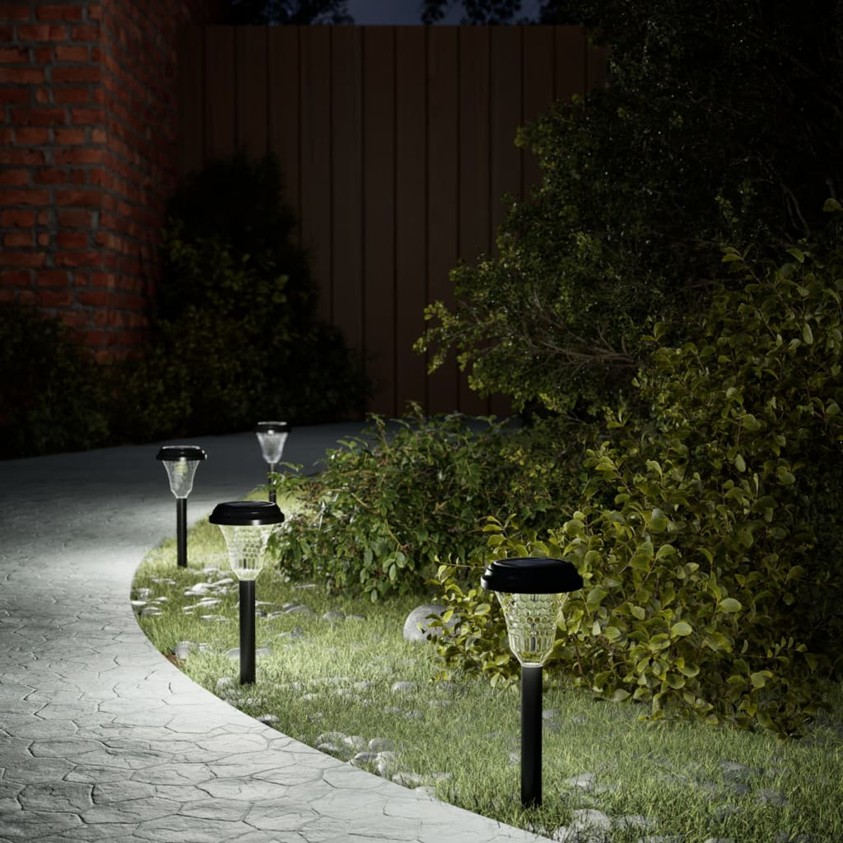 Sentiero illuminato da luci solari da giardino di notte. Cespugli verdi e muro di mattoni.