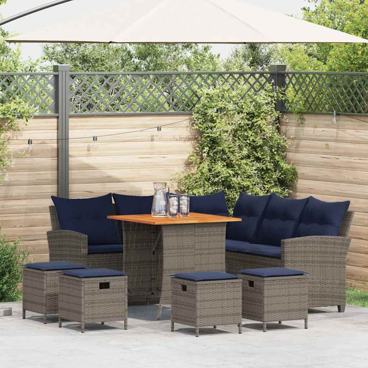 Set di mobili in rattan da esterno con tavolo, divano e sgabelli, cuscini blu, sfondo recinzione in legno.
