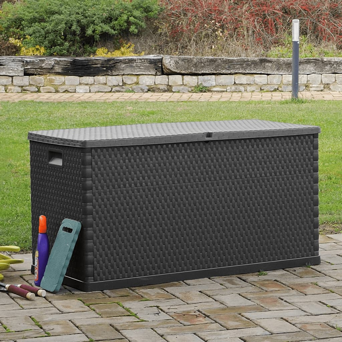 Schwarze Outdoor-Aufbewahrungsbox auf einer Backsteinterrasse, mit Gartengeräten sichtbar.