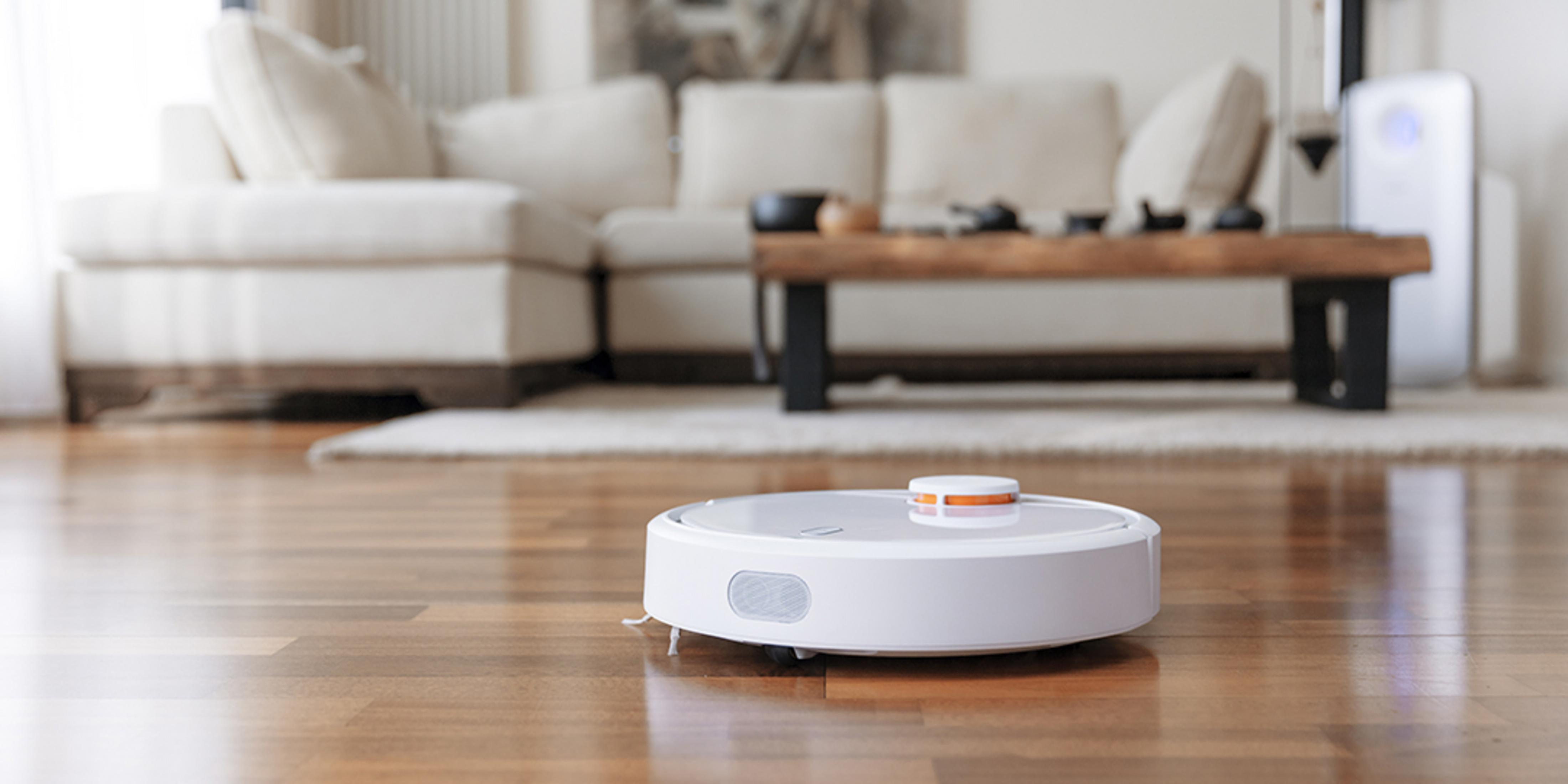 Weißer Roboter-Staubsauger reinigt Holzboden. Wohnzimmer mit Couch im Hintergrund.