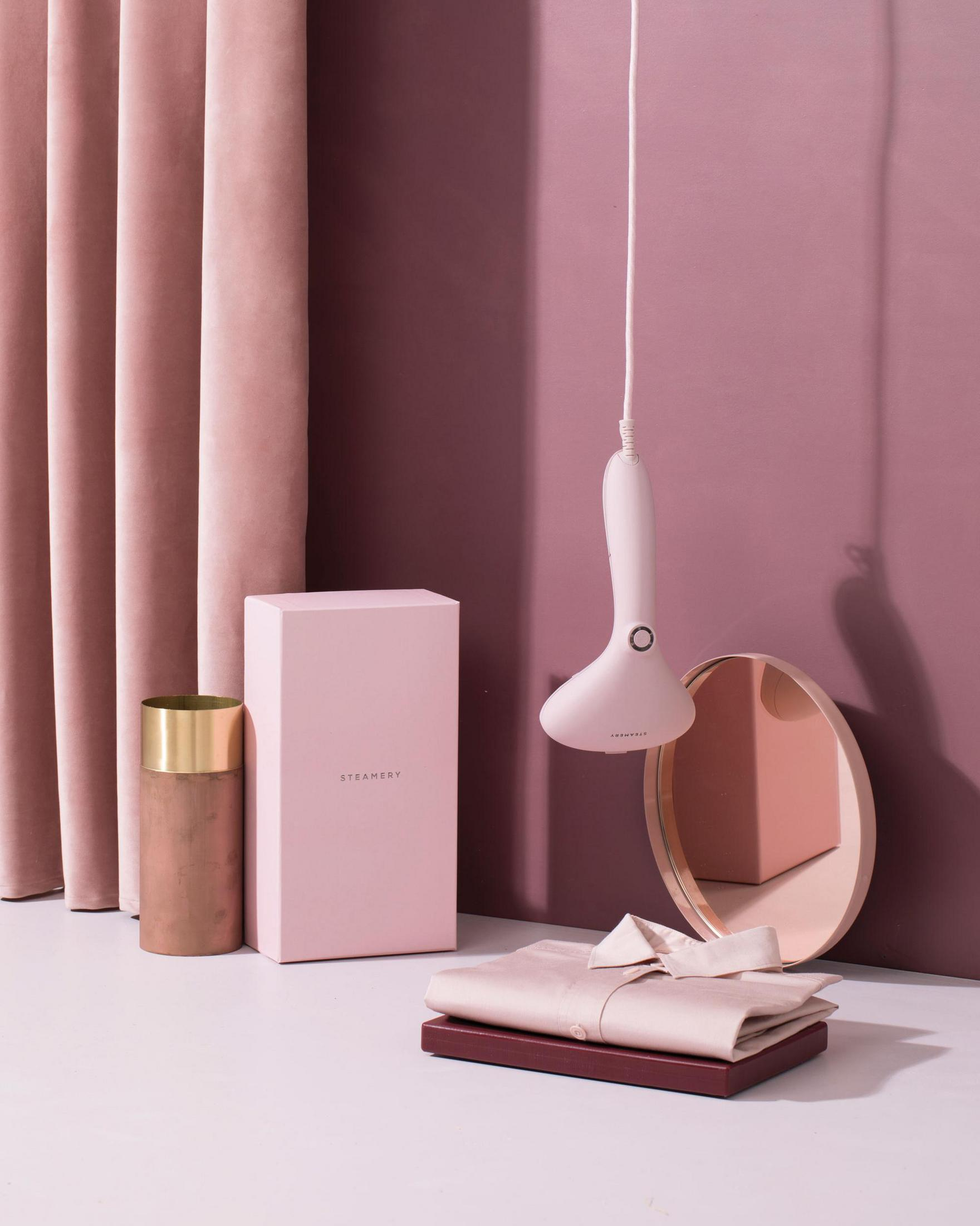 Ein pinker Steamer, eine Box und ein Spiegel stehen auf einer weißen Oberfläche vor pinkem Hintergrund.