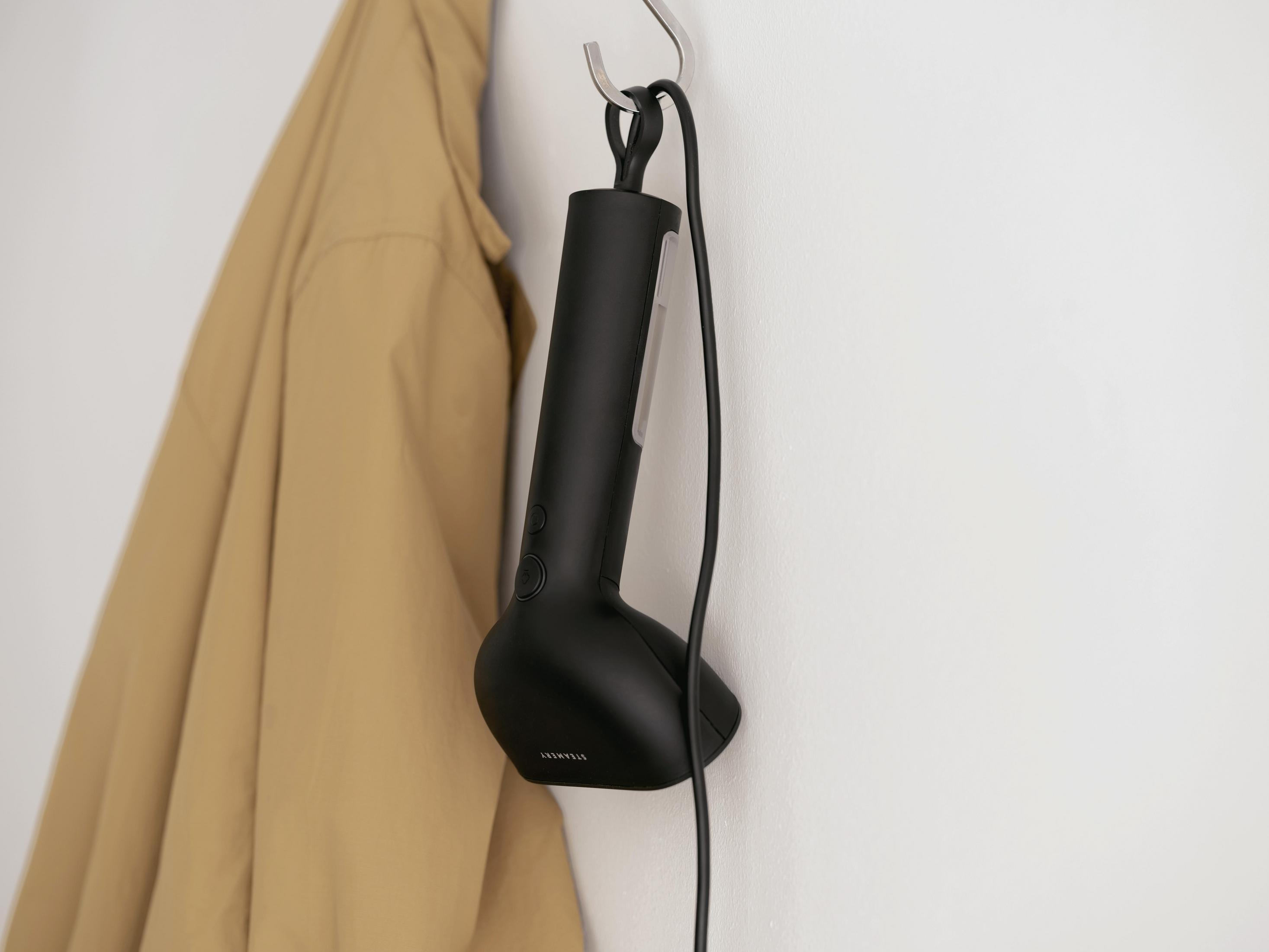 Ein schwarzer Handdampfer hängt an einem Haken neben einer beigen Jacke.