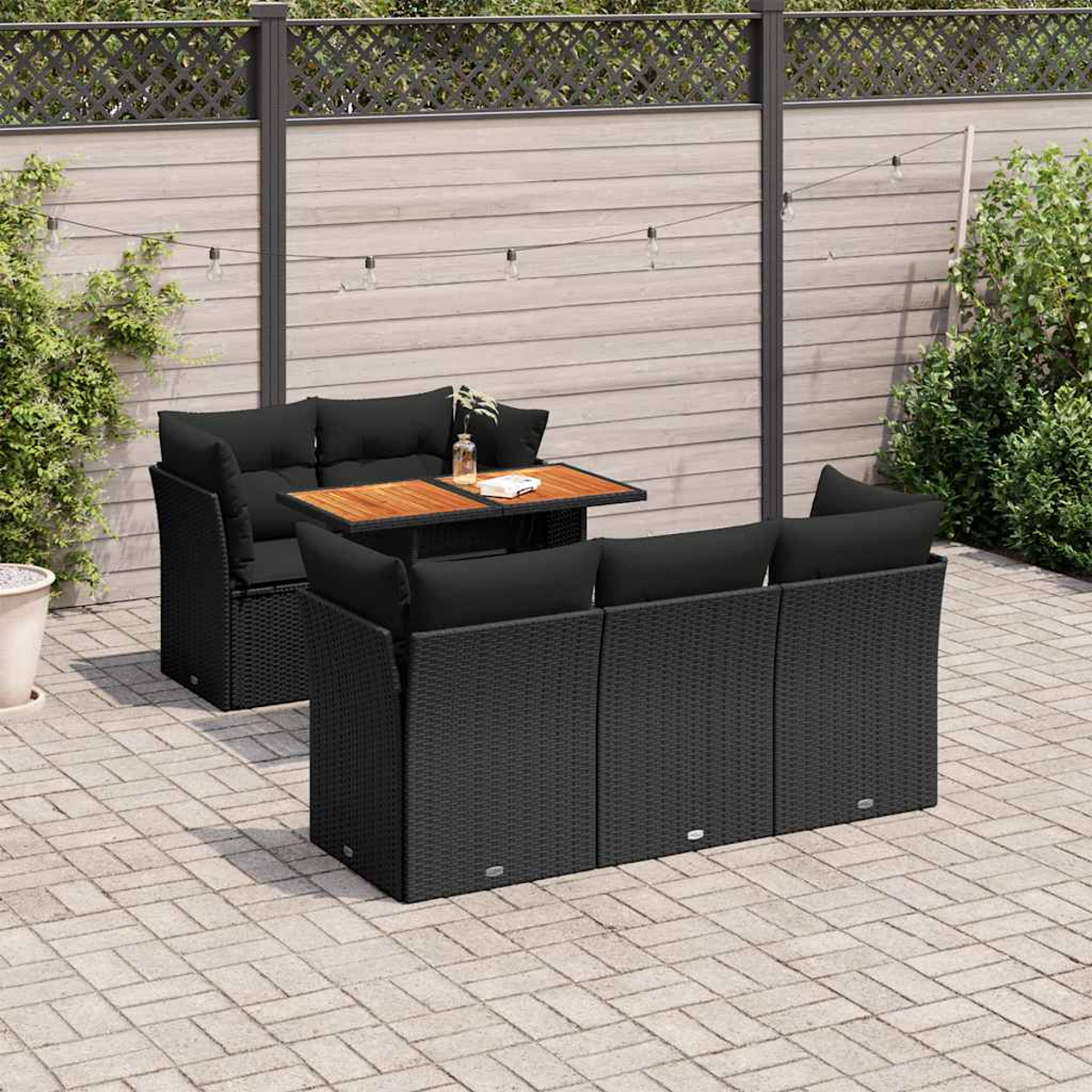 Set patio esterno con cuscini neri, tavolo e piante, contro una recinzione in legno.