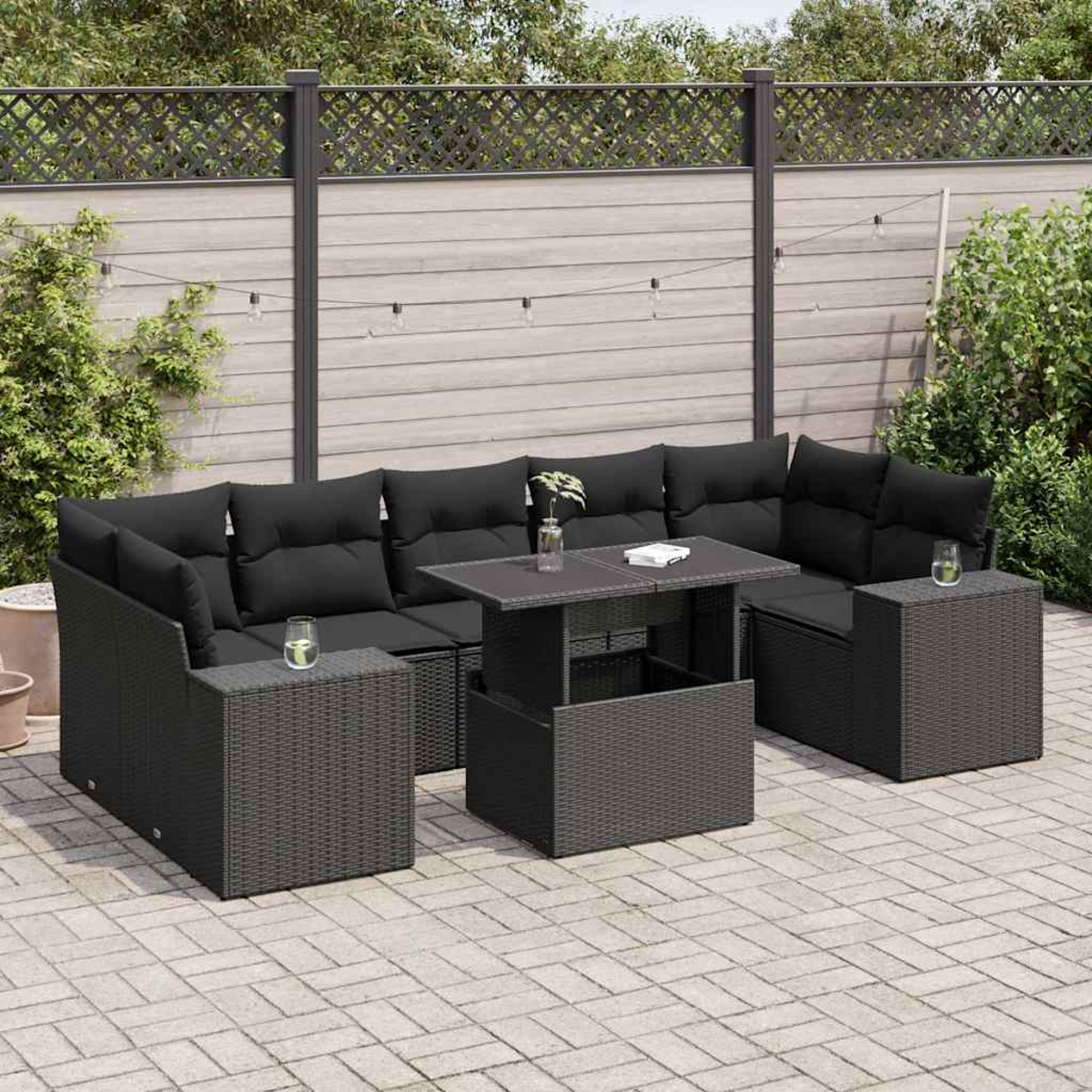 Set divano in rattan nero con tavolo. Vetro, cuscini e piante su un patio in mattoni, con recinzione in legno.