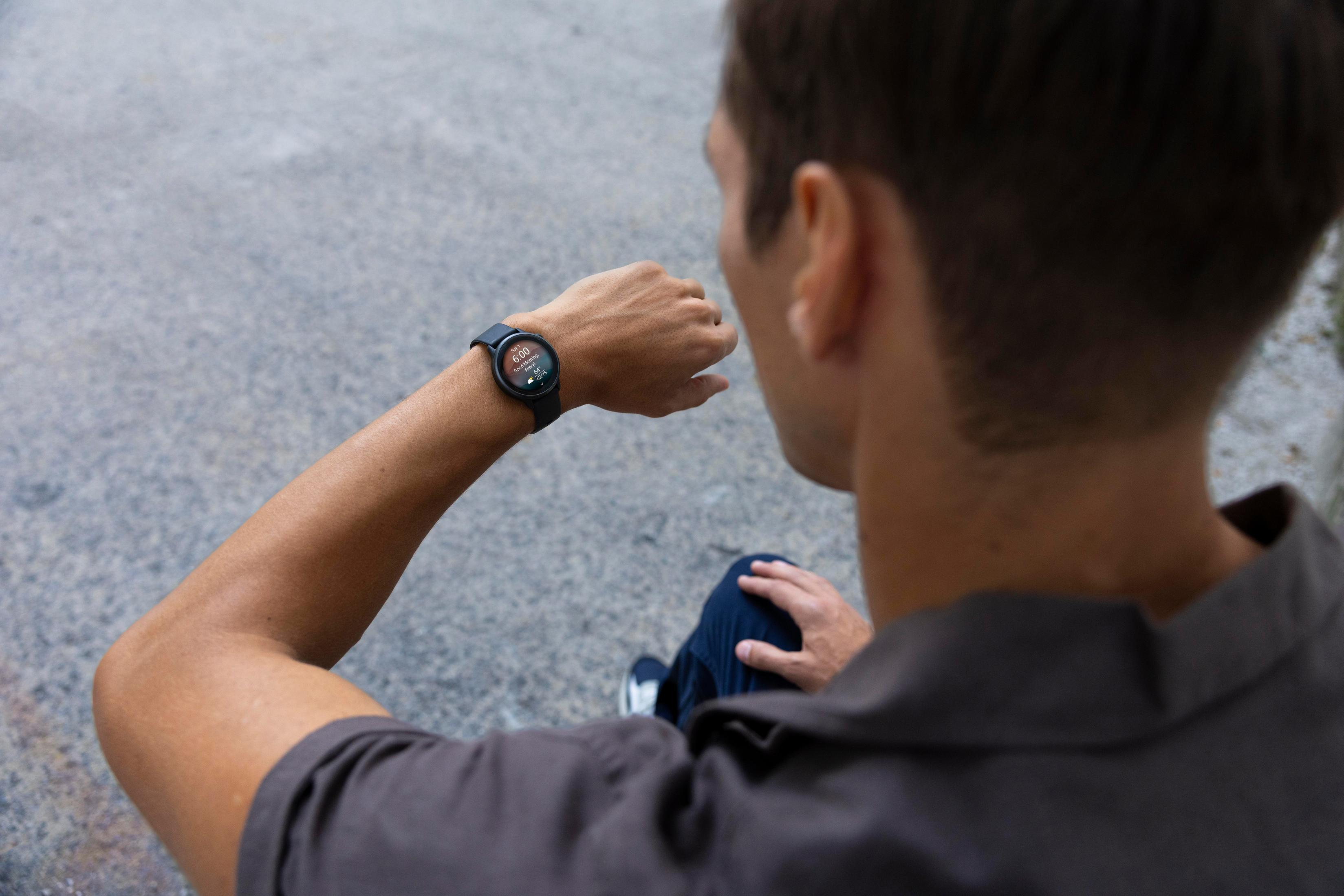 Osoba patrzy na smartwatch. Czas to 6:00. Osoba ma szarą koszulę i siedzi na betonie.