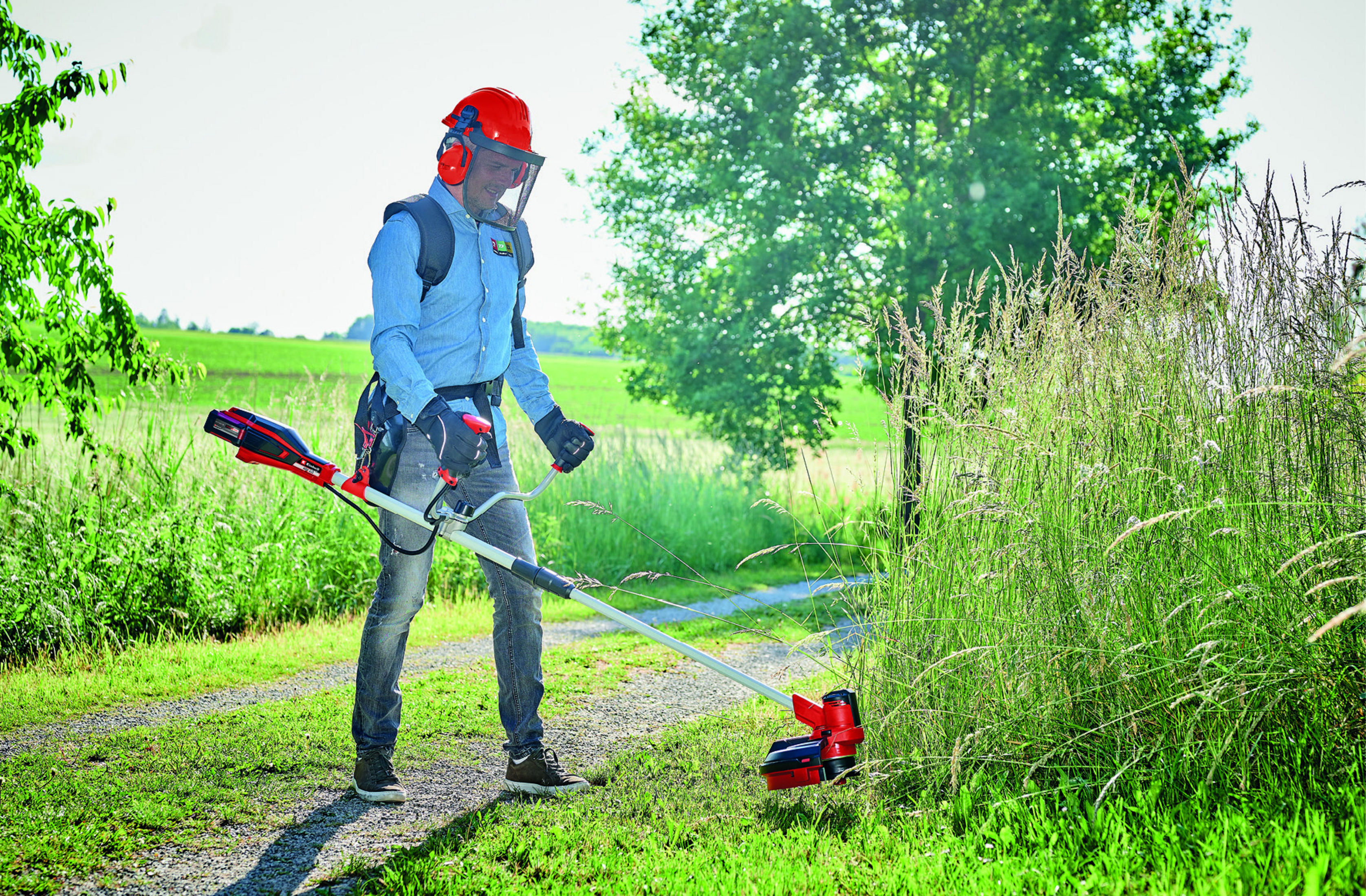 Mann in Schutzkleidung benutzt einen Rasentrimmer auf einem Feld. Grünes Gras, Baum und Himmel im Hintergrund.
