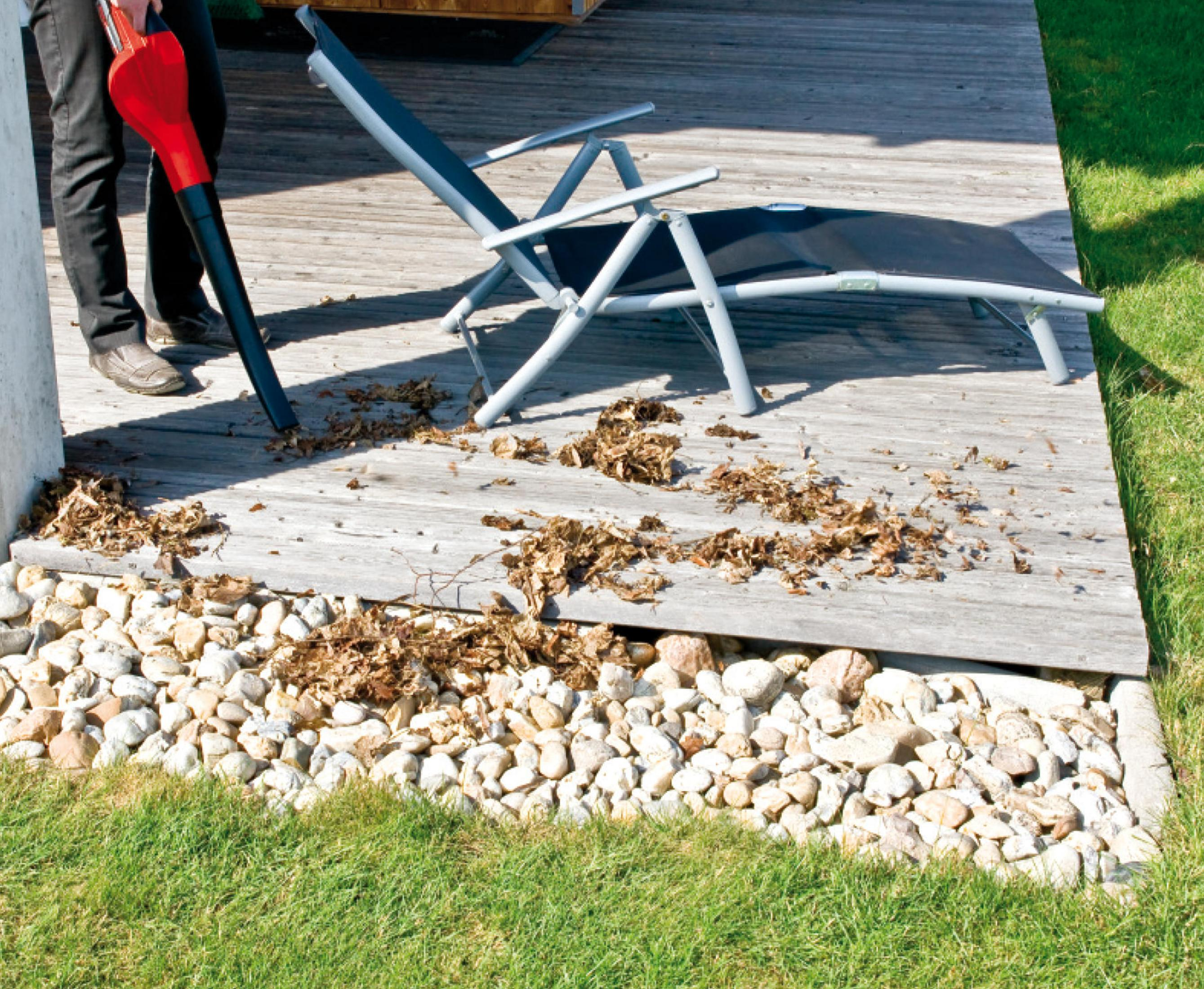 Osoba używa czerwonej dmuchawy do liści do usuwania liści z drewnianego tarasu, obok leżaka.