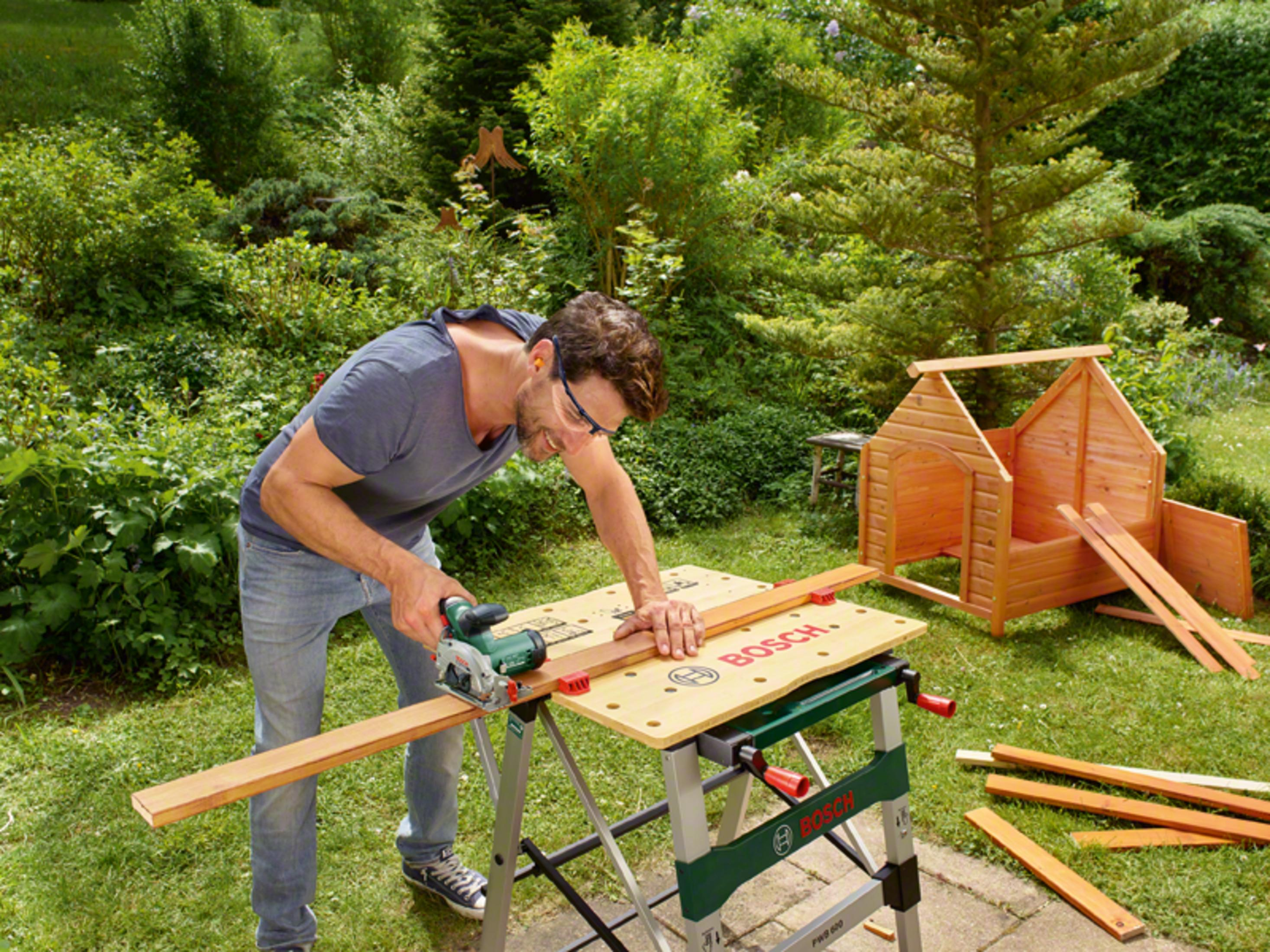 Mężczyzna tnie drewno piłą tarczową na stole warsztatowym. W tle drewniany domek do zabawy.