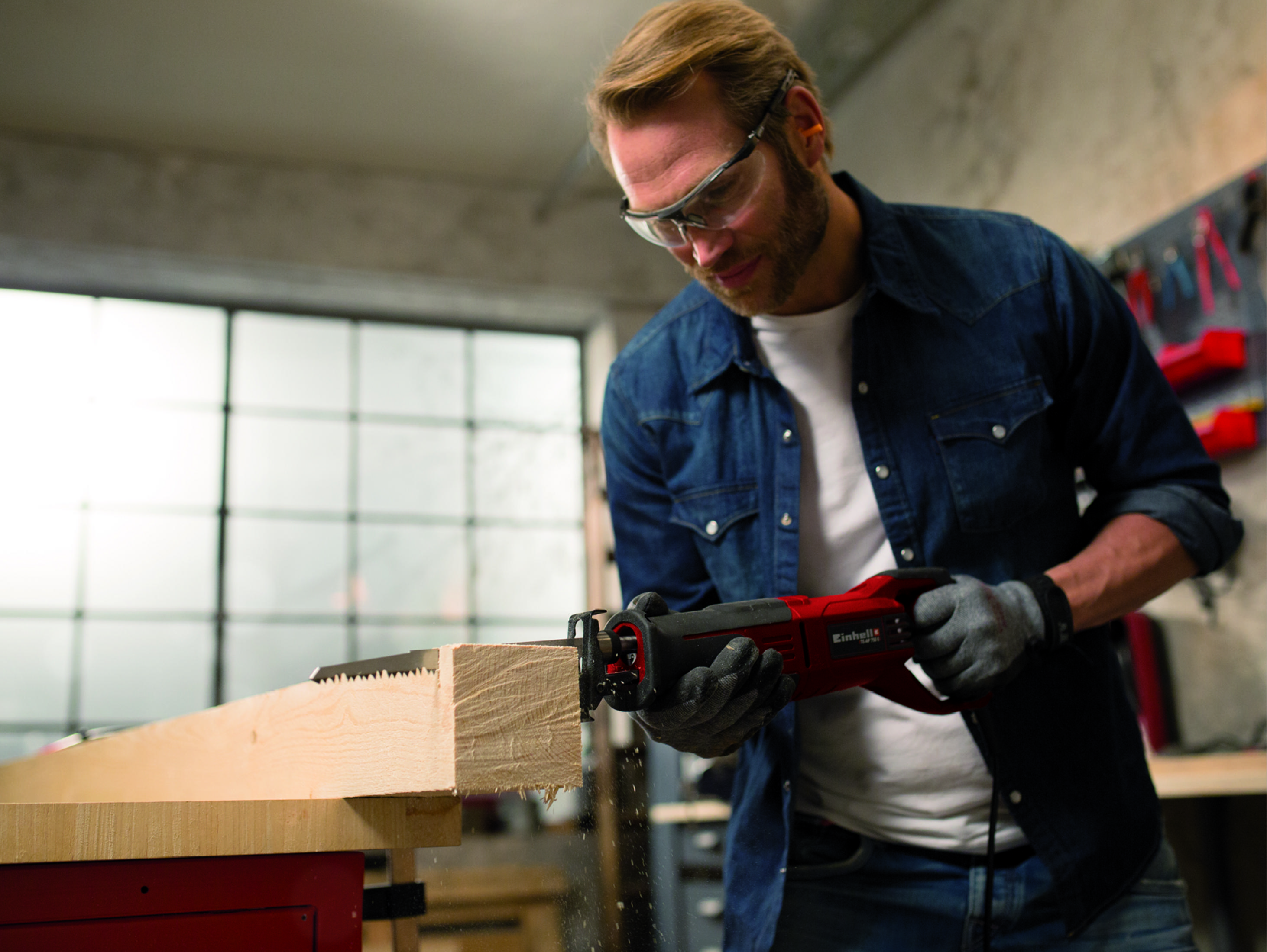 Mężczyzna w okularach ochronnych używa czerwonej piły szablastej do cięcia drewna.