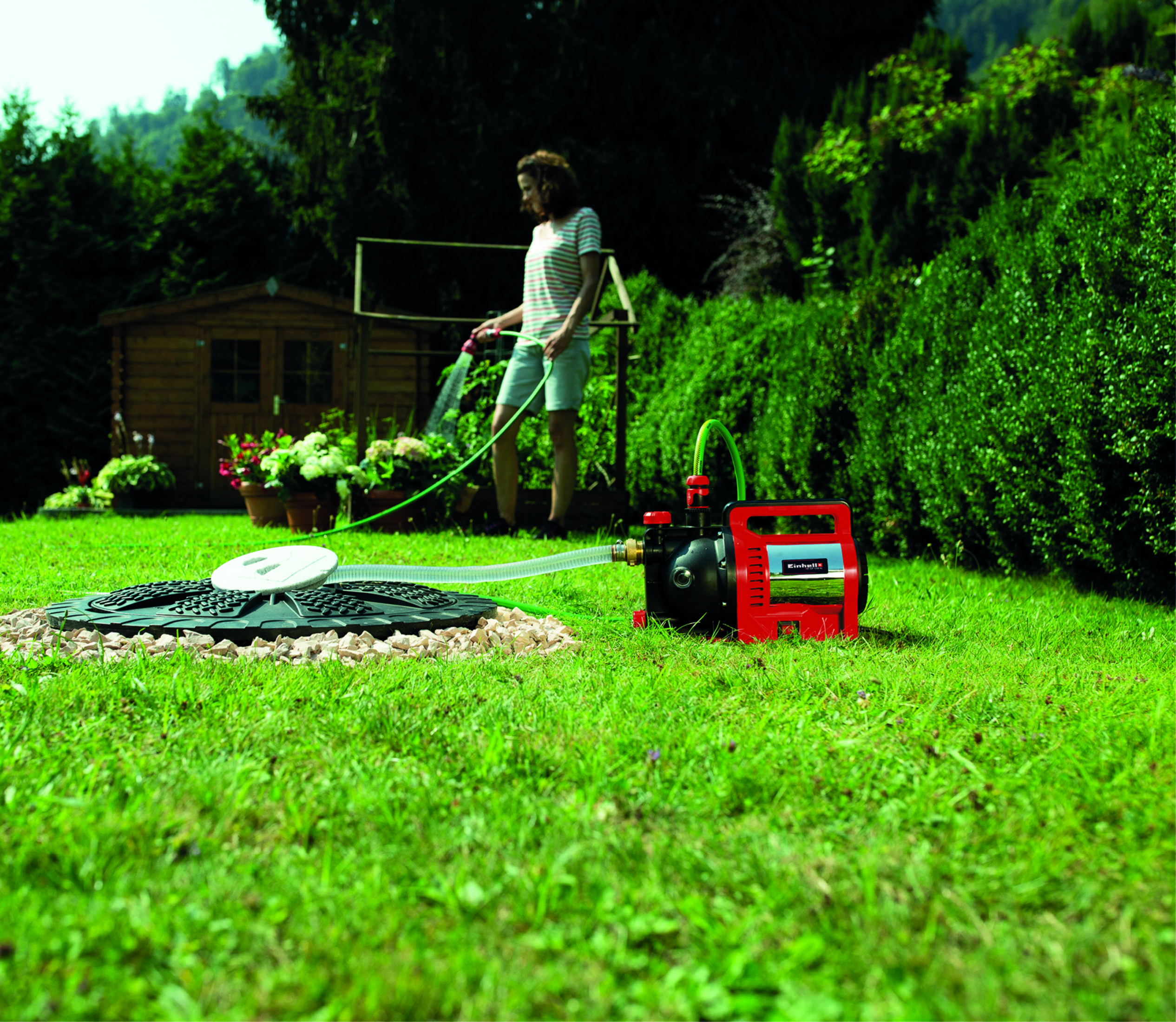 Eine Frau gießt einen Garten mit einem Schlauch. Eine Pumpe und ein Schuppen sind im Hintergrund sichtbar.