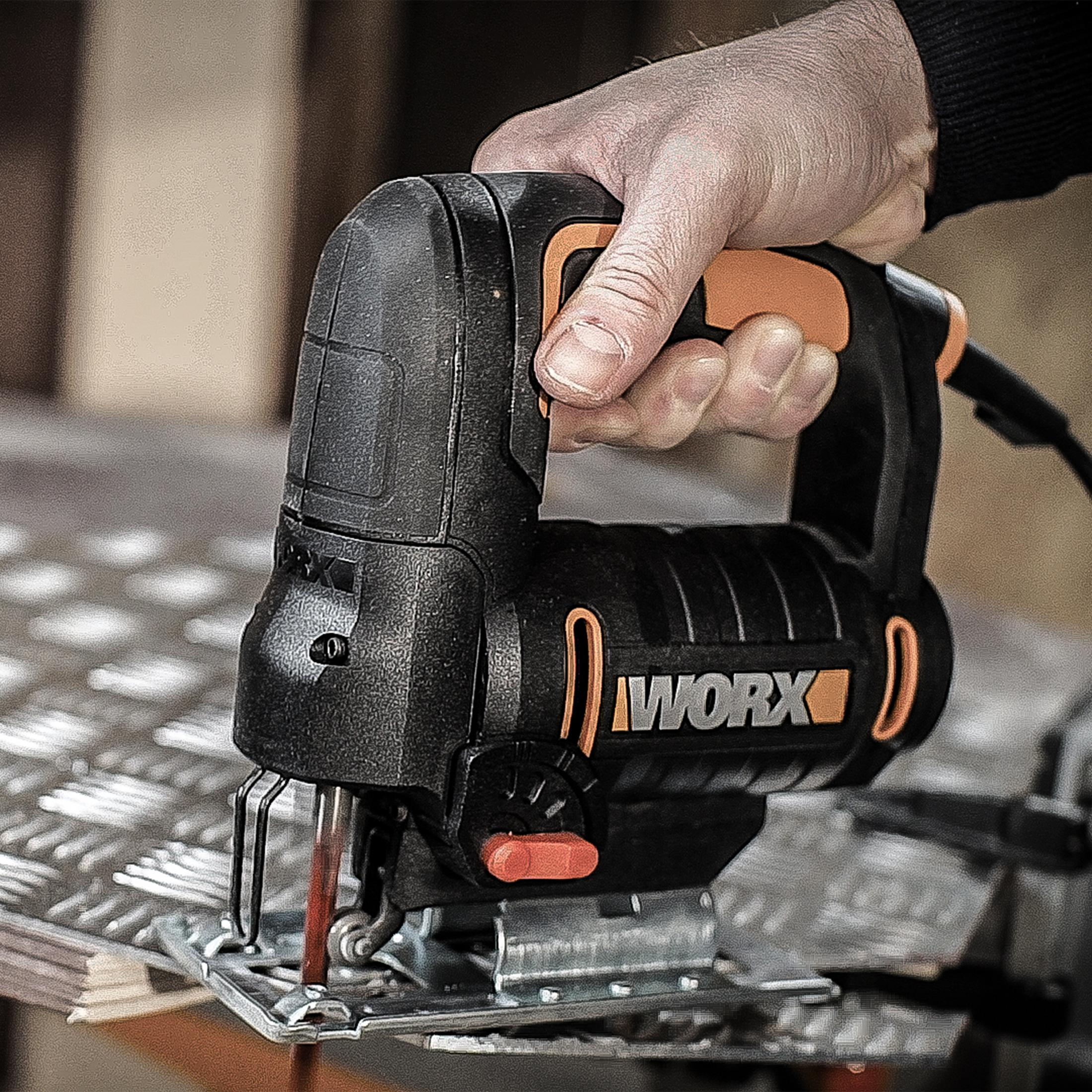Eine Hand hält eine schwarz-orangefarbene WORX Stichsäge. Das Sägeblatt befindet sich in der Nähe einer Metalloberfläche. Der Hintergrund ist unscharf.