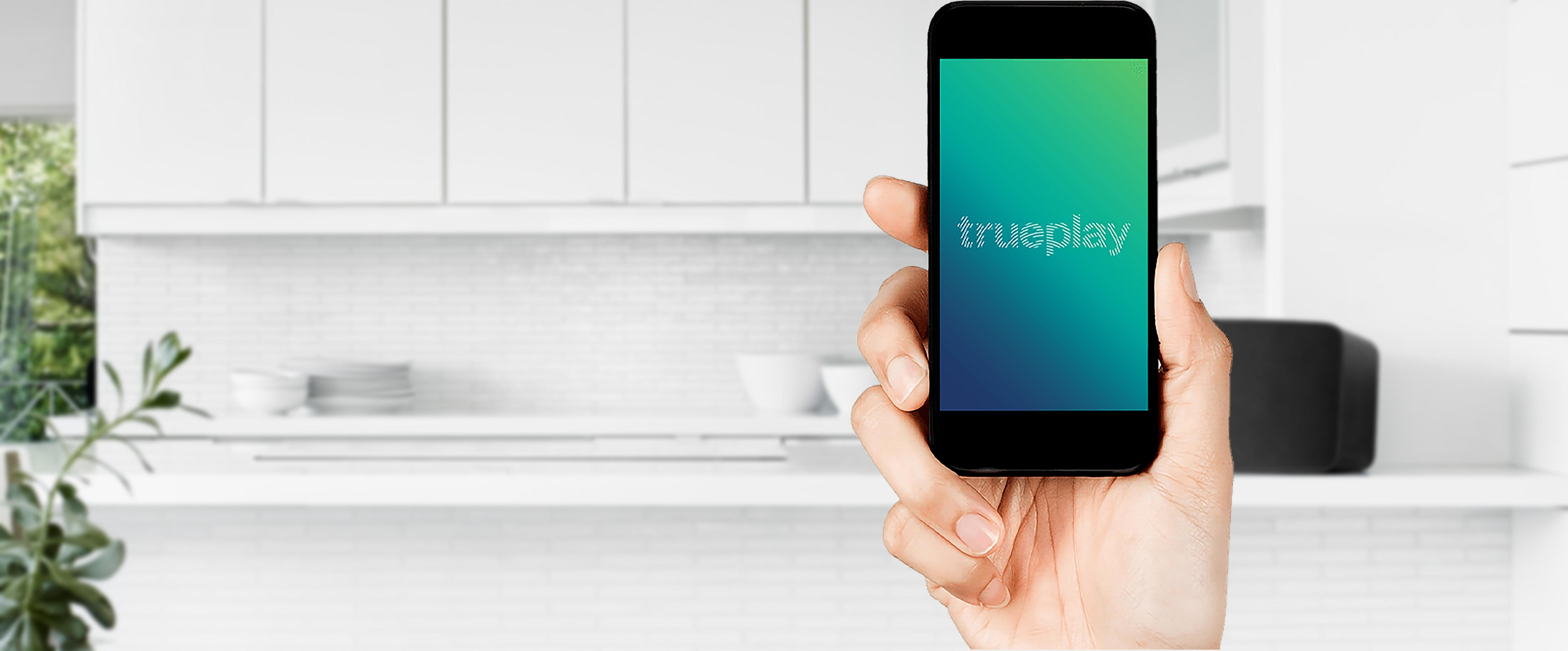 Beyaz bir mutfağın önünde Trueplay uygulaması açık olan bir telefon tutan bir el. Arkada bir hoparlör var.