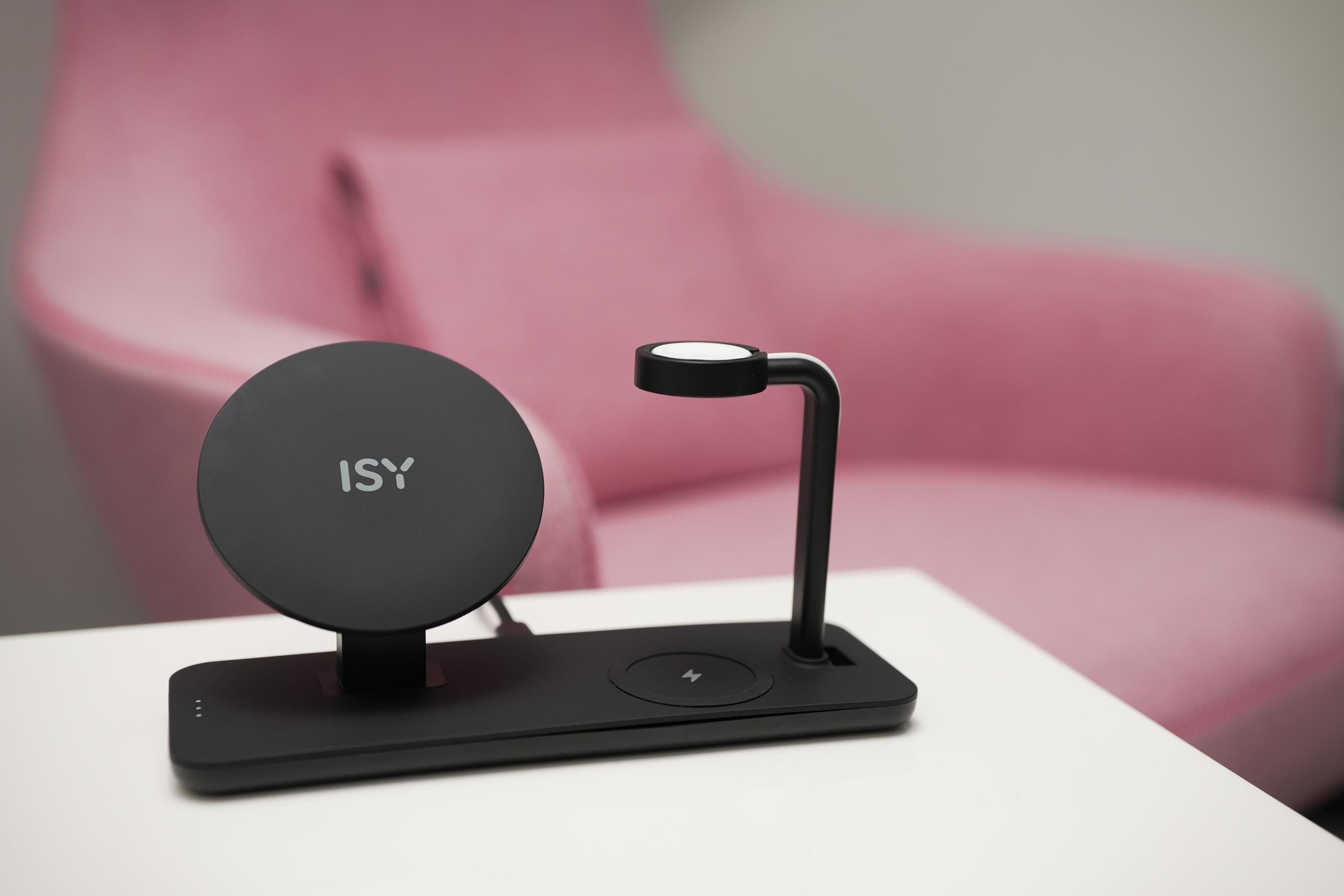 Schwarze kabellose Ladestation mit 'ISY'-Logo auf einem Tisch und einem rosa Sessel im Hintergrund.