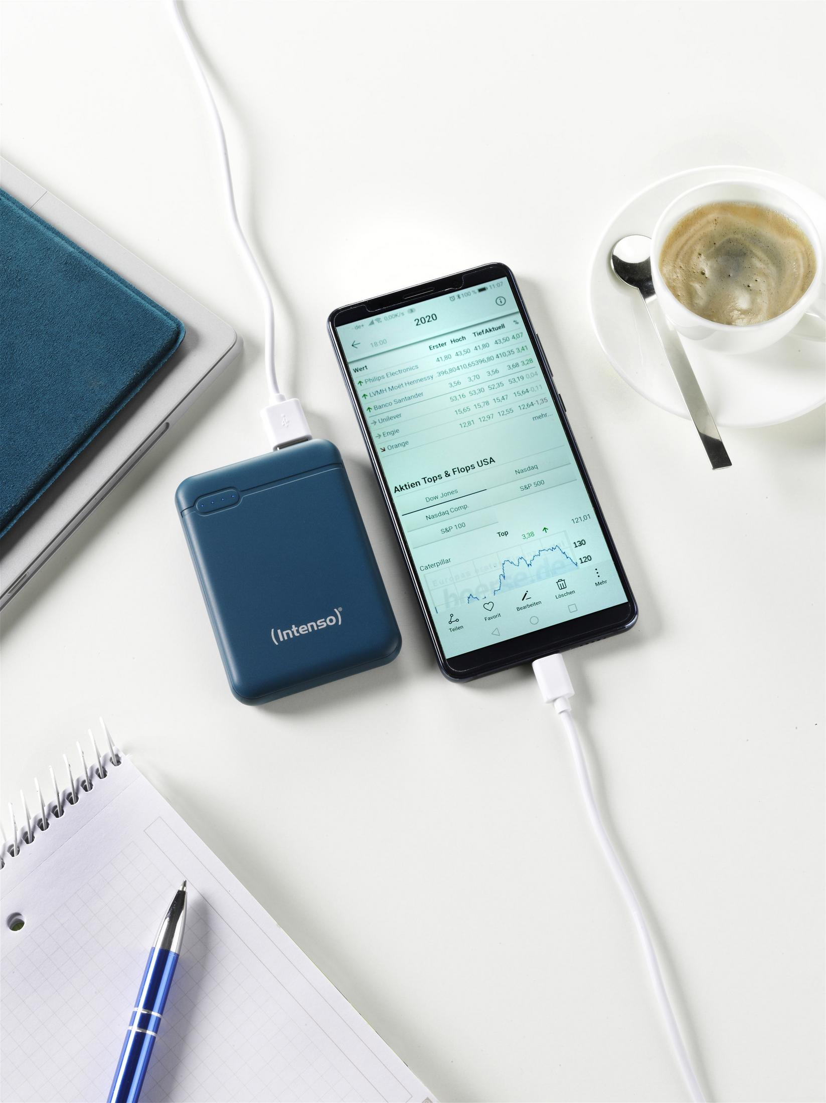 Telefon, das von einer blauen 'Intenso' Powerbank geladen wird, mit Laptop, Stift und Kaffeetasse auf dem Tisch.
