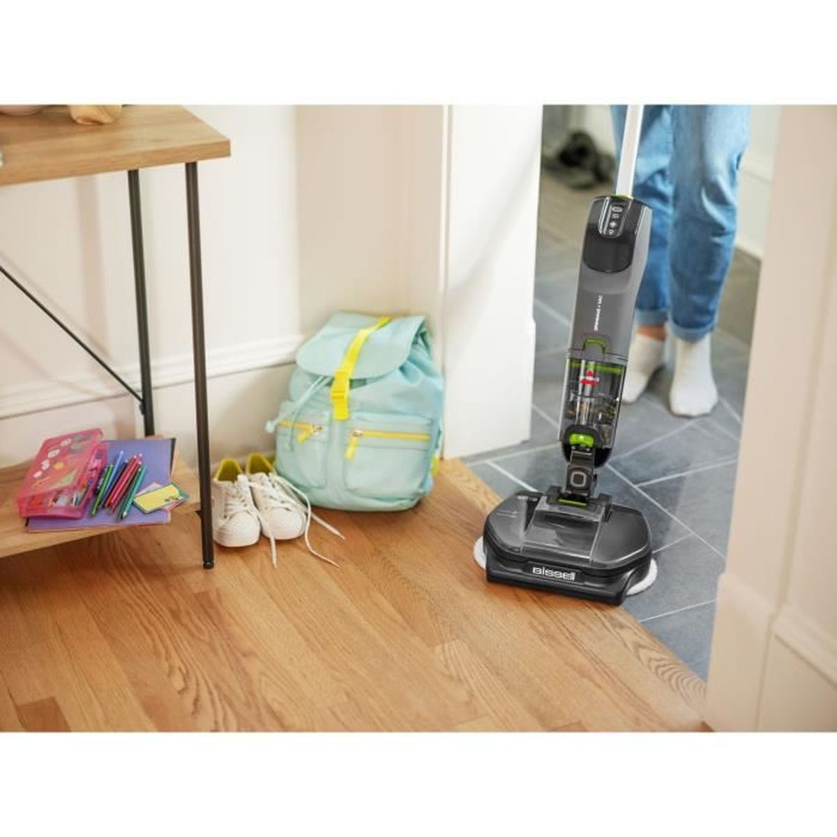 Una persona limpia un piso de madera con una aspiradora. Una mochila y zapatos están en el fondo.