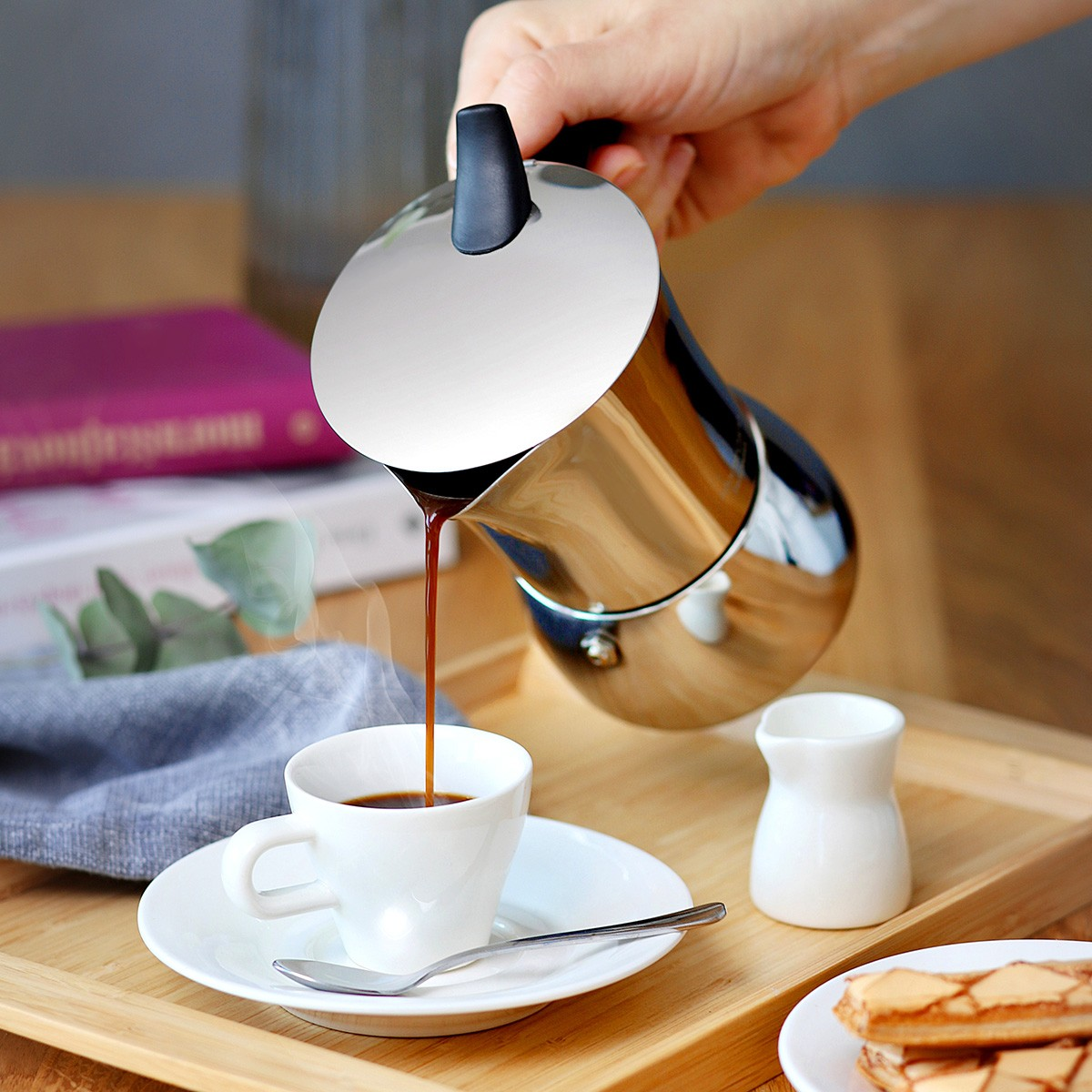 Eine Person gießt Kaffee aus einer glänzenden silbernen Kanne in eine weiße Tasse auf einer Untertasse. Daneben steht ein kleiner Sahnebereiter.