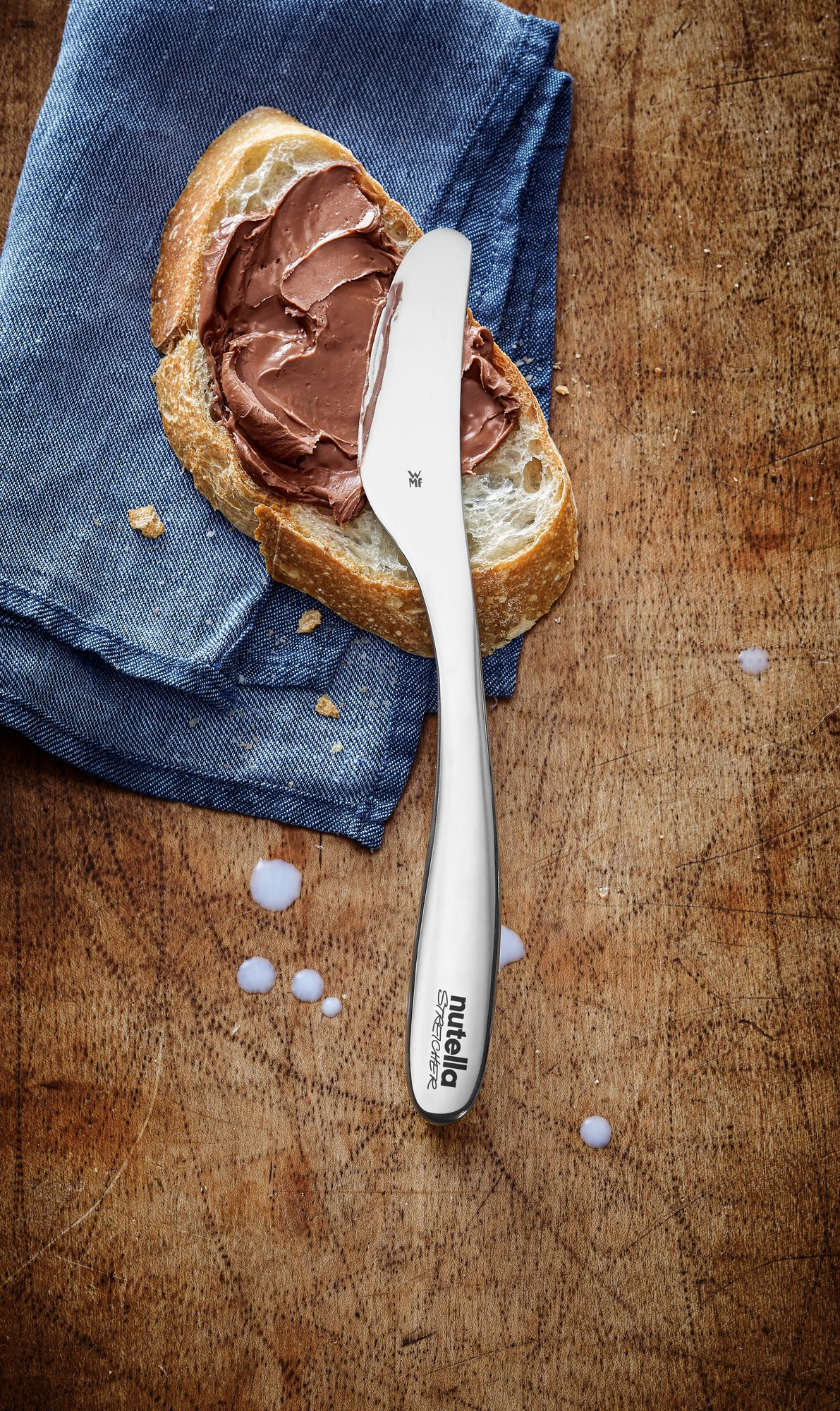 Kawałek chleba z Nutellą i nożem na drewnianej desce. Widoczna niebieska szmatka.