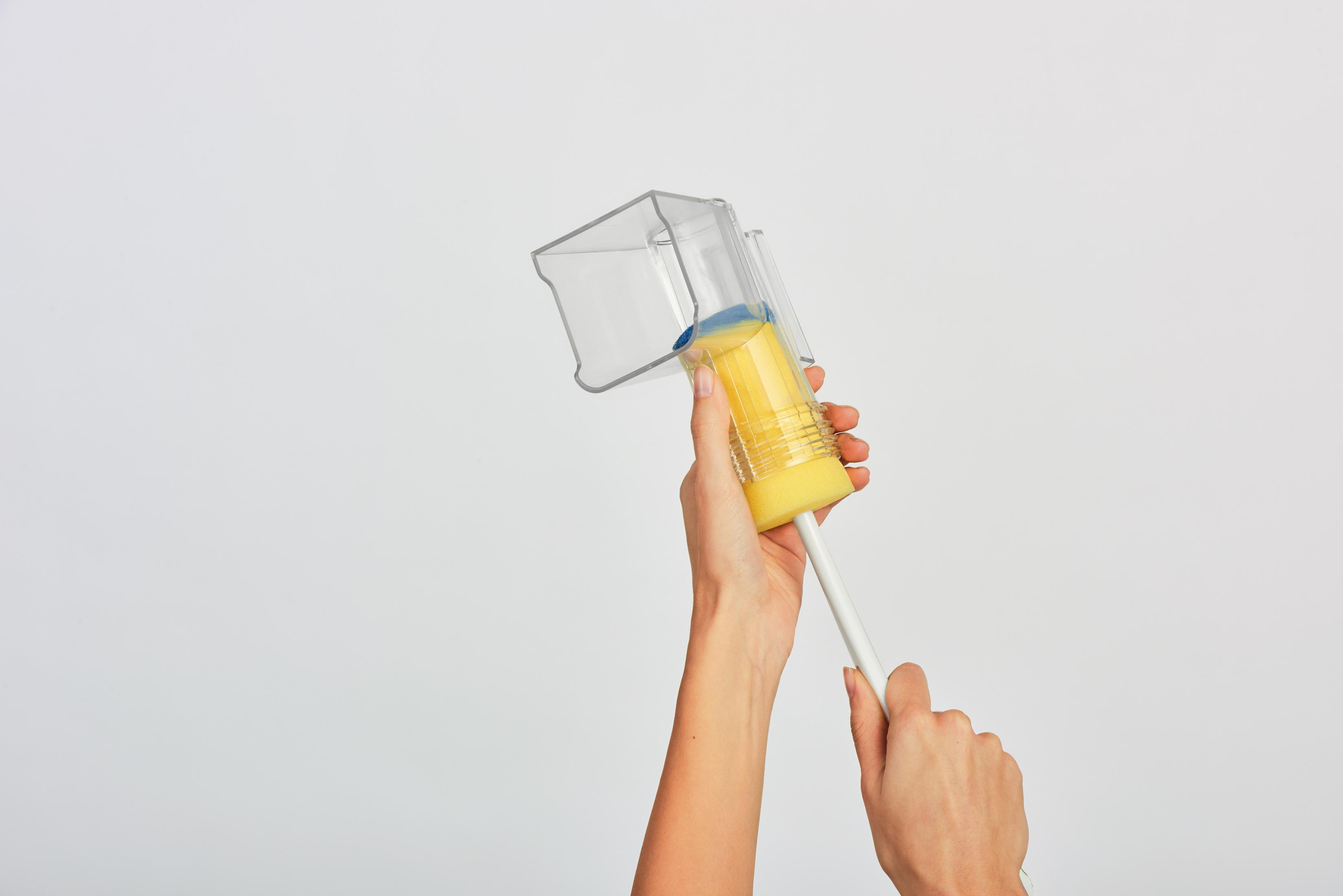 Ręce trzymają narzędzie do czyszczenia gąbką. Ma żółtą gąbkę, przezroczysty pojemnik i biały uchwyt.