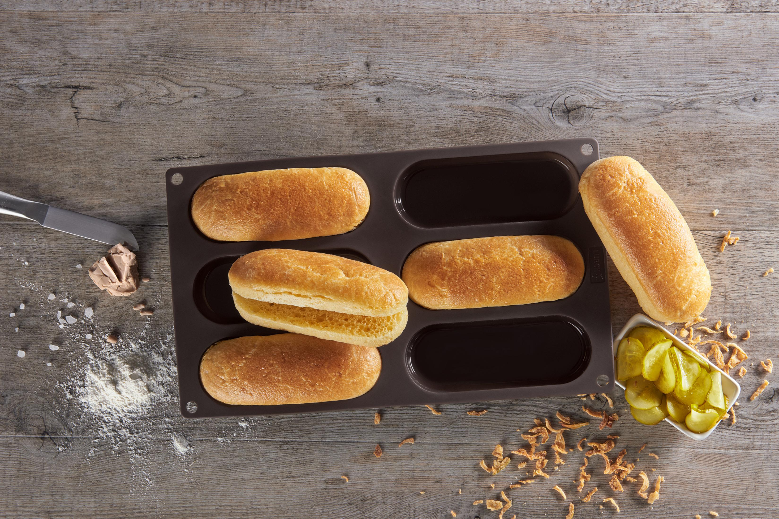 Sechs gebackene Hotdog-Brötchen in einer Schale. Eines ist offen, andere Artikel sichtbar. Holzoberfläche.