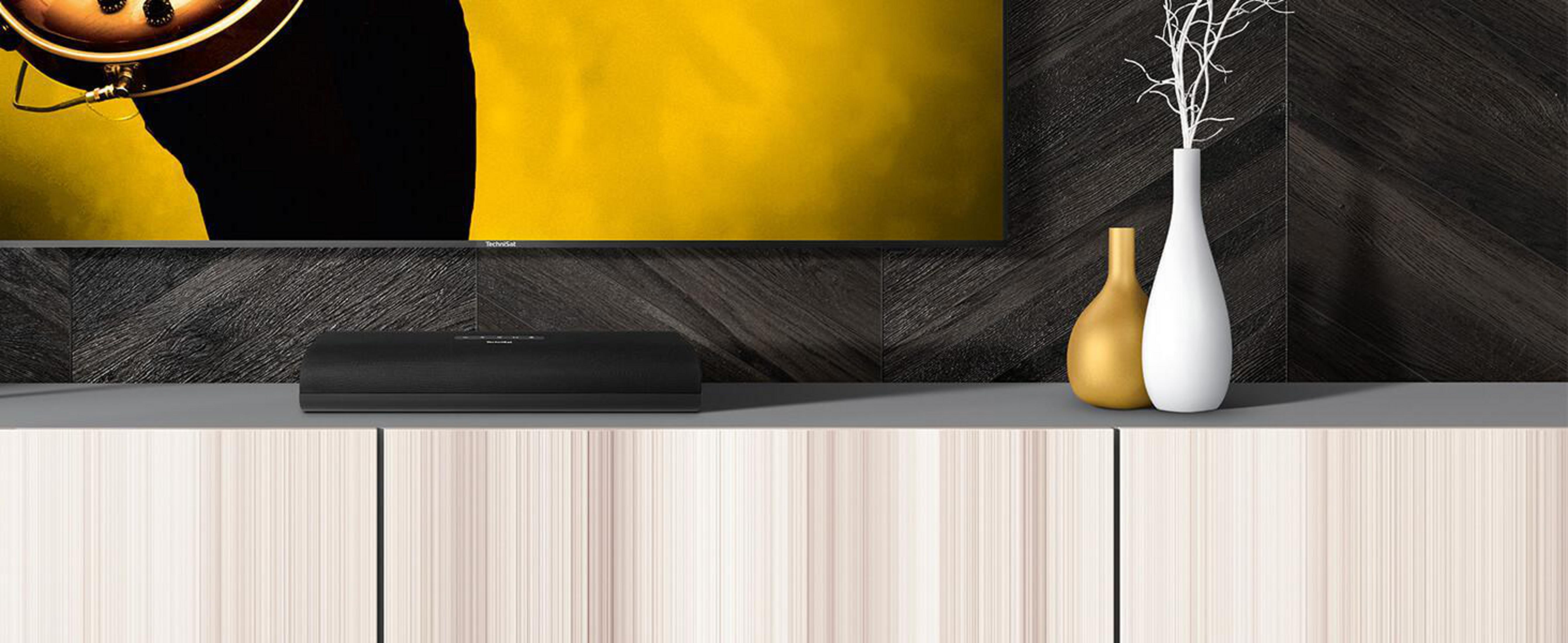 Soundbar na szafce z wazonami na górze. Żółte i białe wazony, ciemne panele drewniane.