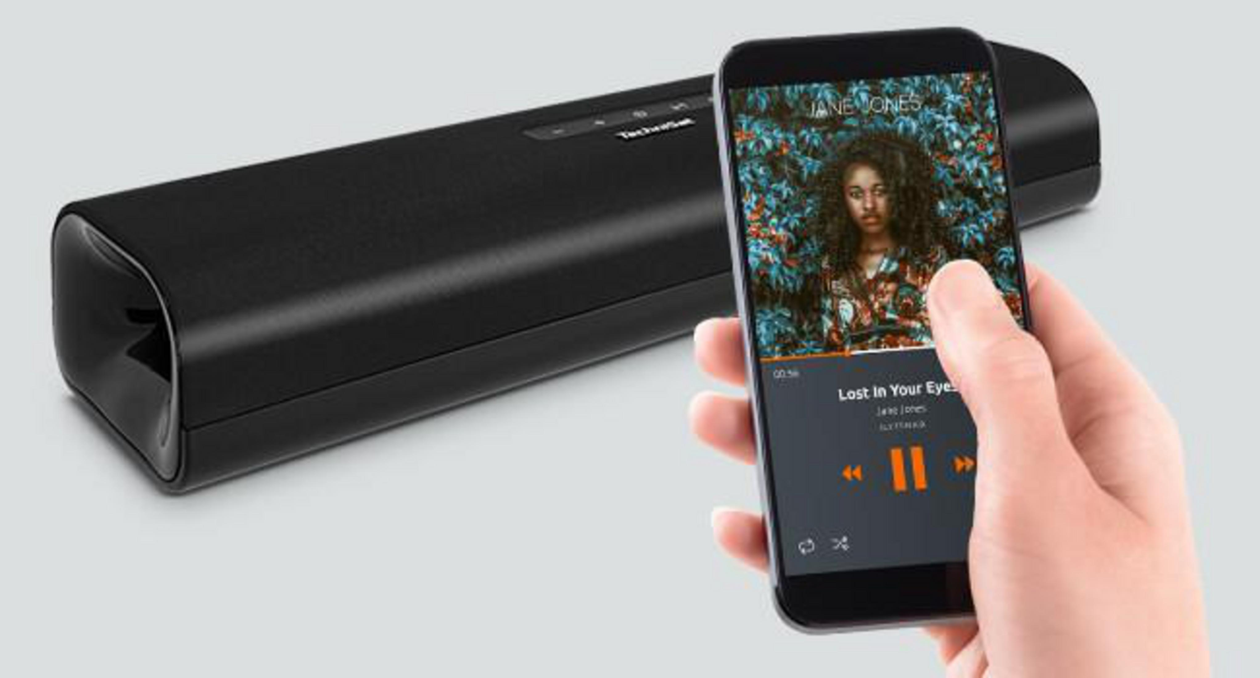 Dłoń trzymająca smartfon z muzyką, obok czarnego soundbaru. Jasne tło.