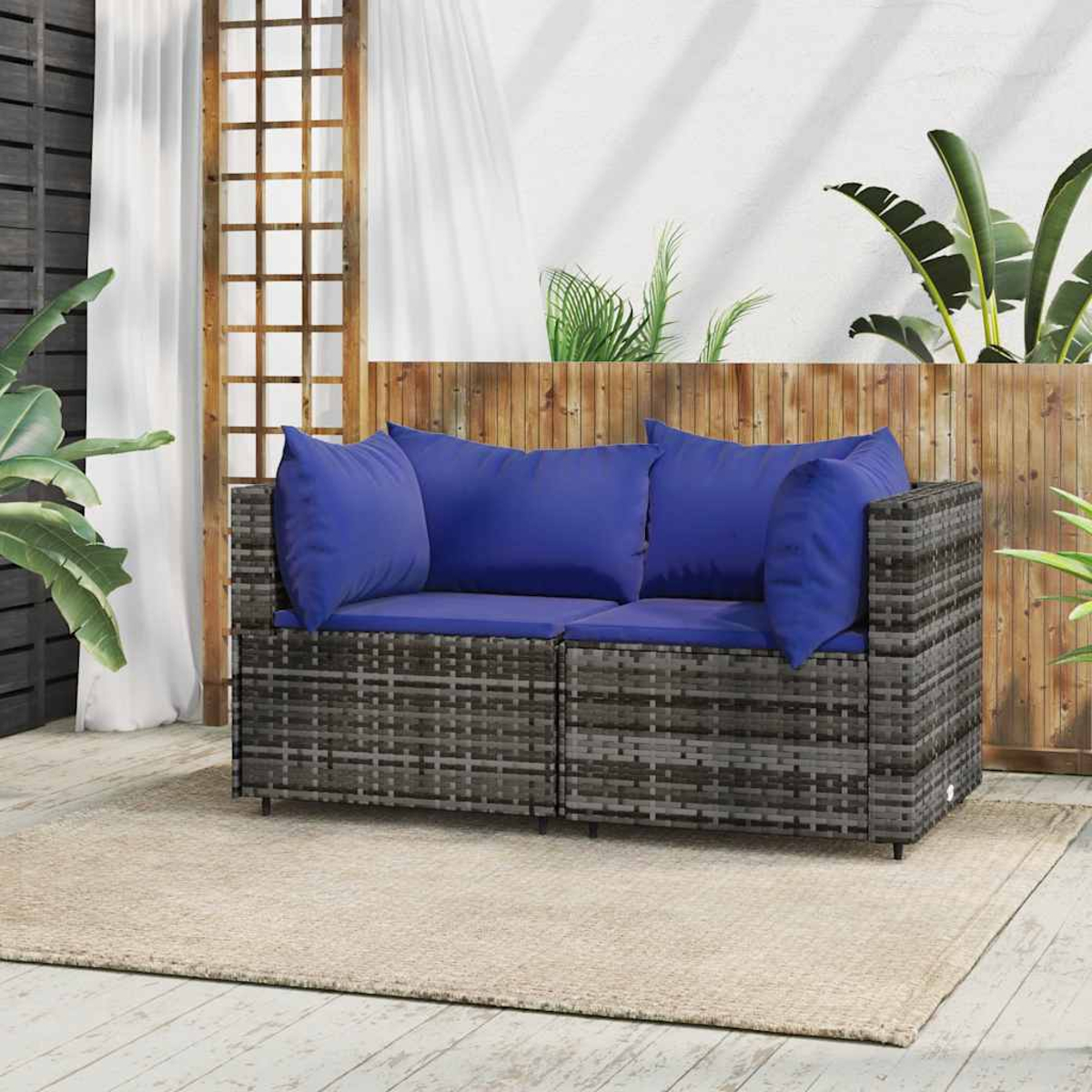 Rattan-Sofa im Freien mit blauen Kissen. Beiger Teppich, Holzzaun. Pflanzen im Hintergrund, sonnige Umgebung.