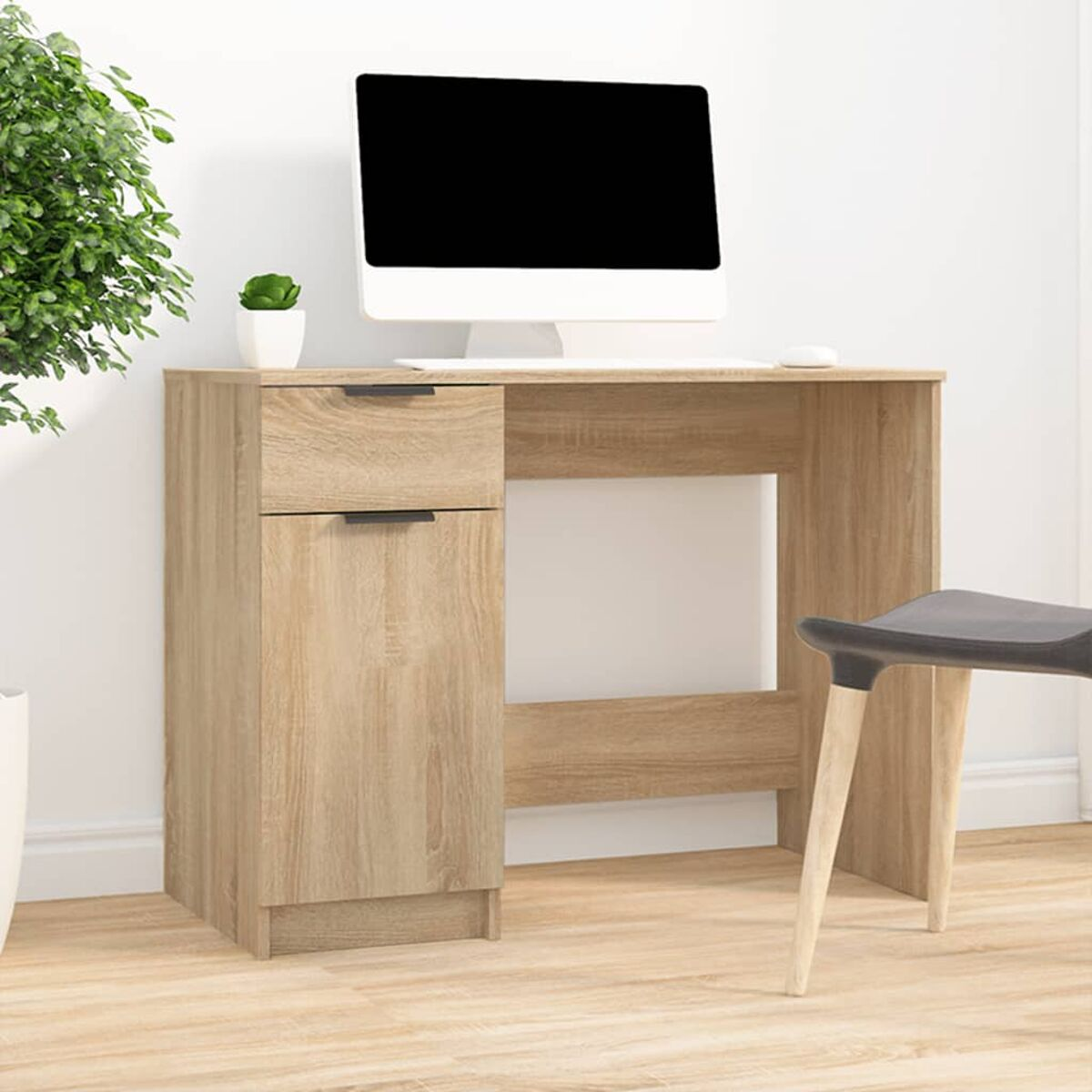 Holzschreibtisch mit Schubladen und Computermonitor. Stuhl und Pflanze daneben. Weiße Wand.