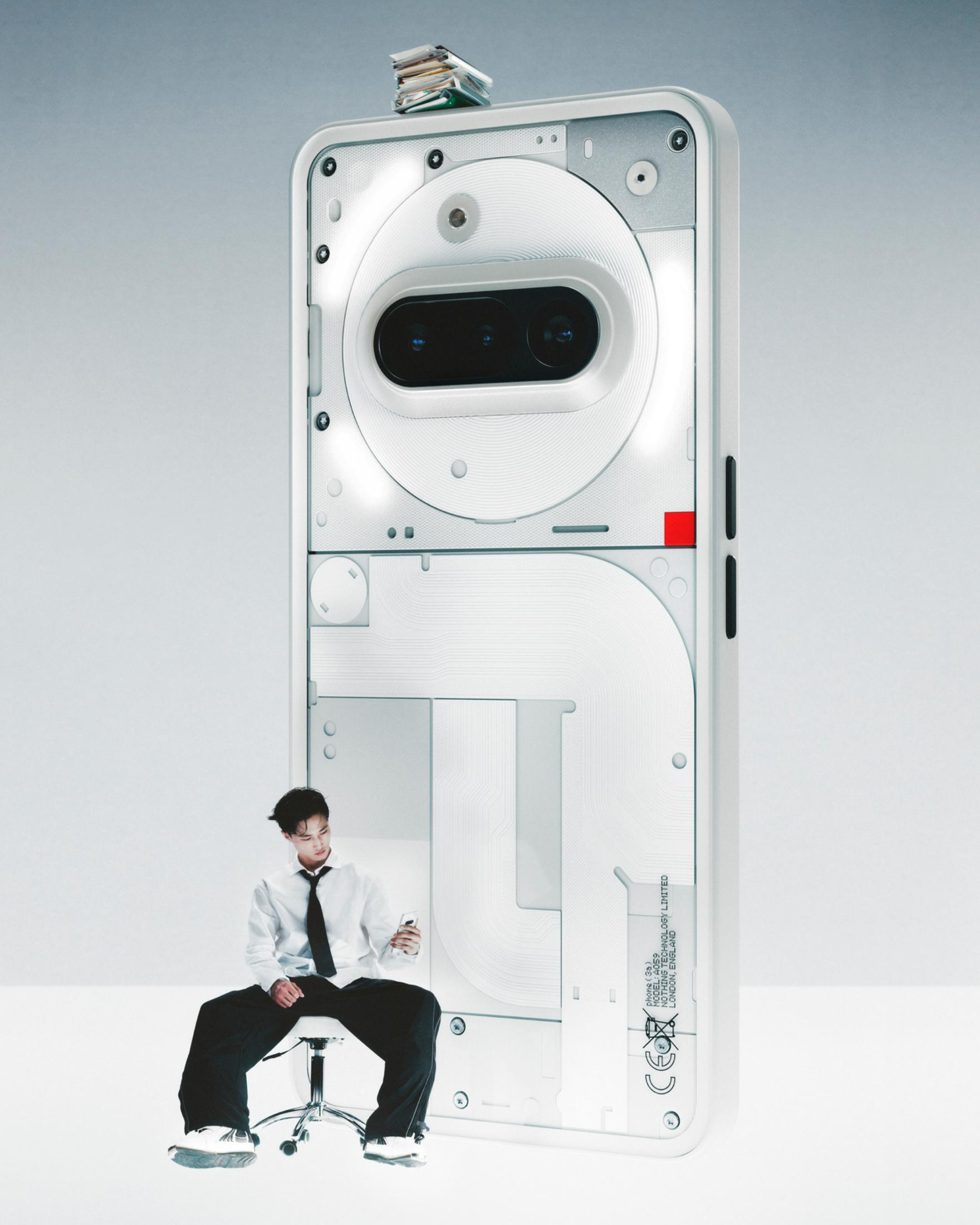 Mężczyzna siedzi obok dużego białego telefonu z przezroczystym tyłem, trzymając mniejszy telefon.