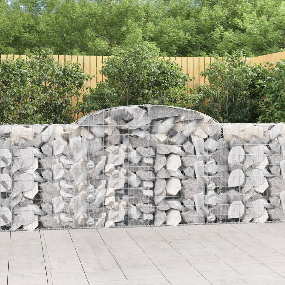 Gabionenwand mit grauen Steinen, vor grünen Büschen und einem Holzzaun.