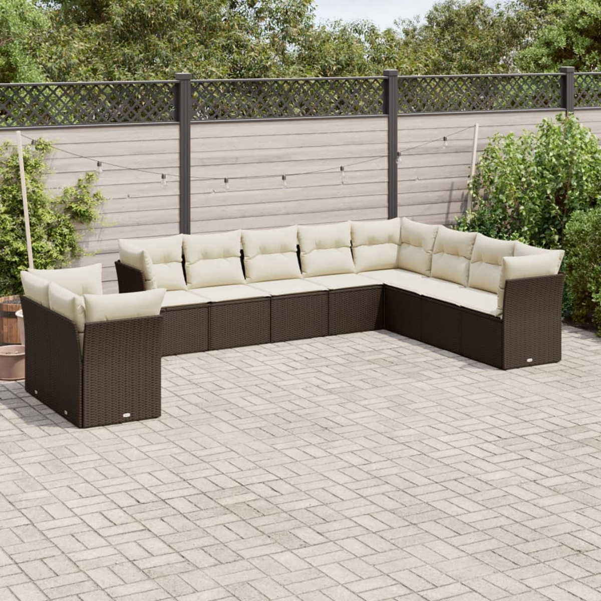 Braunes Outdoor-Ecksofa aus Korbgeflecht mit cremefarbenen Kissen auf einer Ziegelterrasse. Zaun und Grün im Hintergrund.