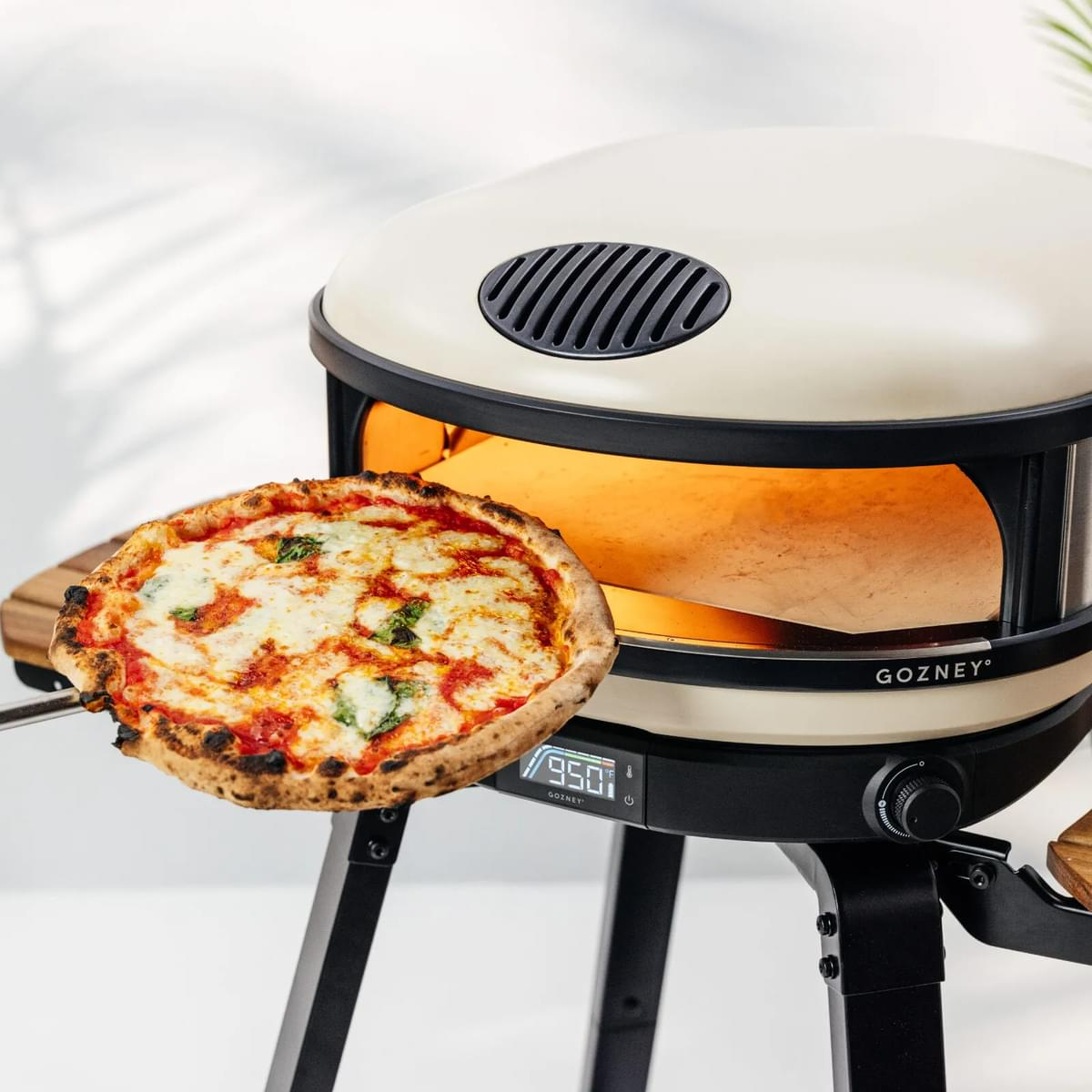 Eine Pizza wird aus einem Pizzaofen genommen. Der Ofen ist cremefarben, auf einem schwarzen Ständer.