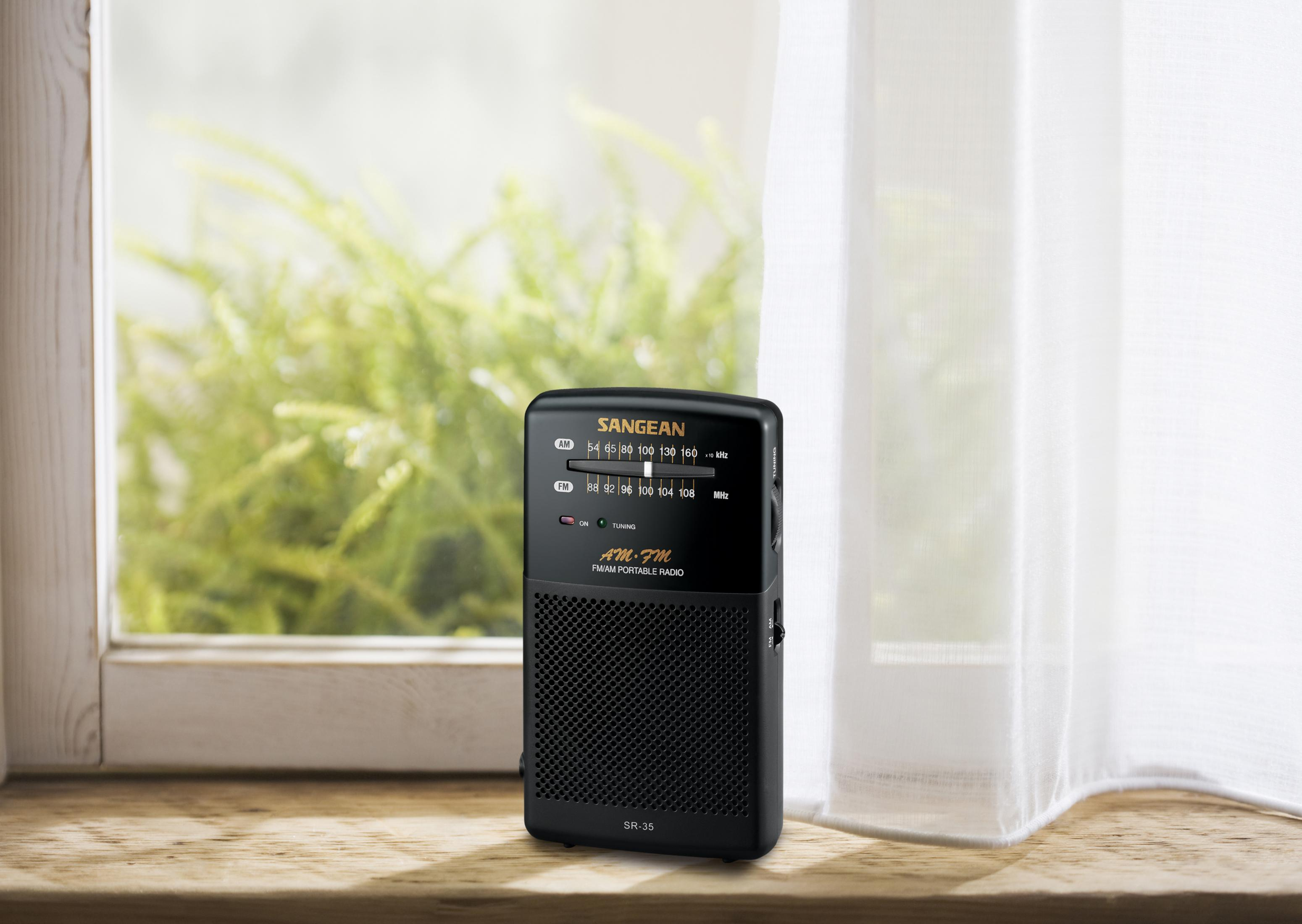 Black Sangean radio on a wooden surface, near a window with plants and a white curtain.