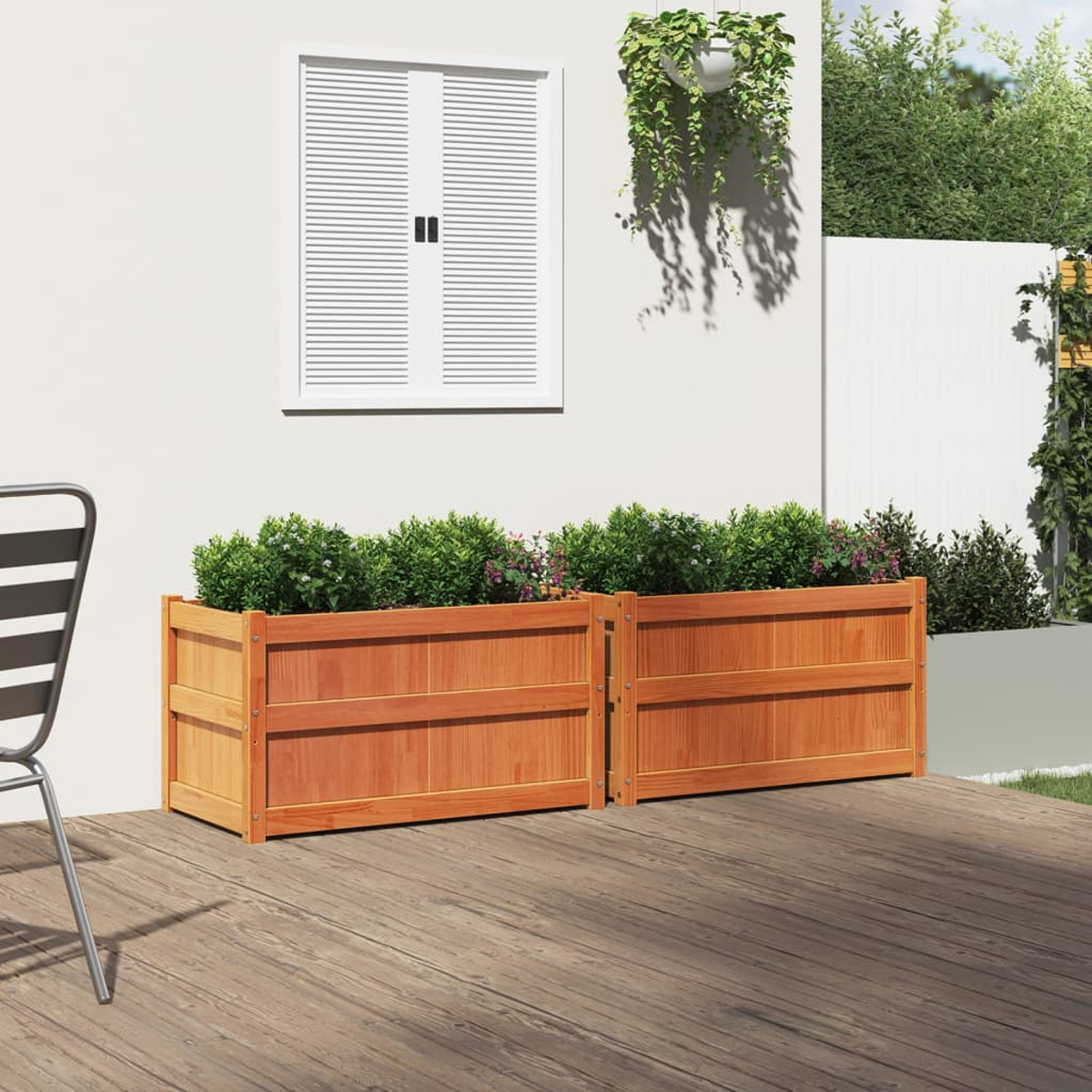 Zwei Holzpflanzkästen mit grünen Pflanzen auf Holzterrasse. Weiße Wand und Fenster im Hintergrund.