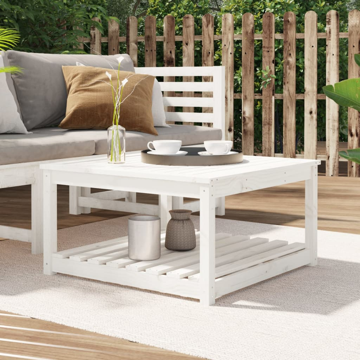 Ein weißer quadratischer Holztisch mit einem unteren Regal. Ein Sofa und Pflanzen sind im Hintergrund.