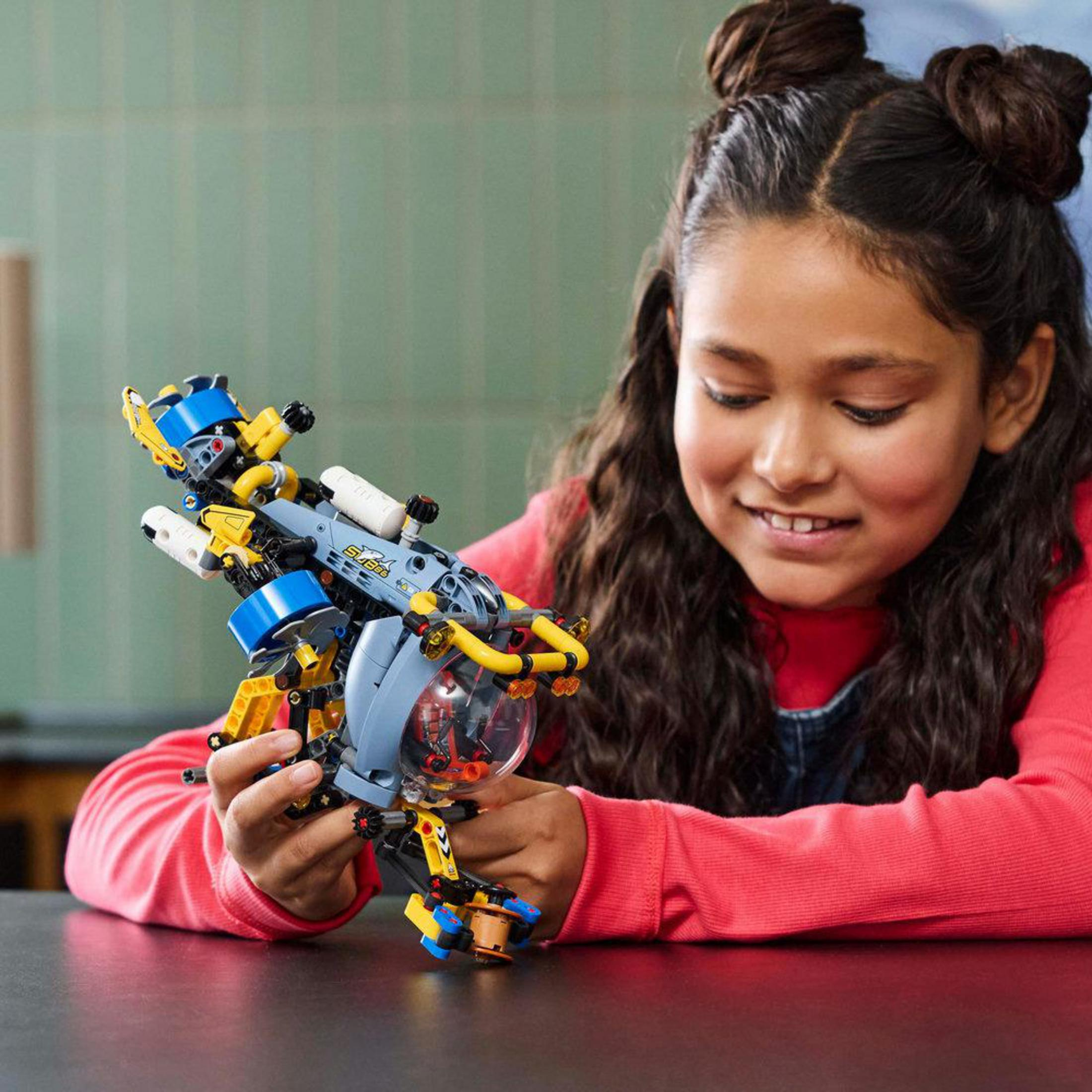 Sarı ve mavi detaylara sahip bir Lego Technic denizaltı modeli tutan gülümseyen bir kız.