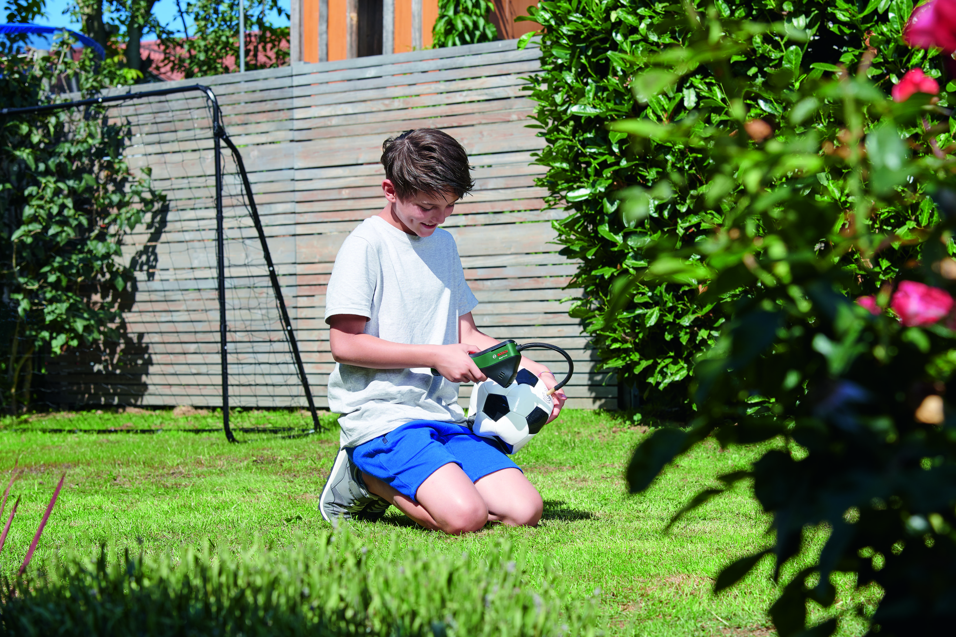 Junge kniet auf Gras, pumpt einen Fußball auf. Garten und Zaun sichtbar.