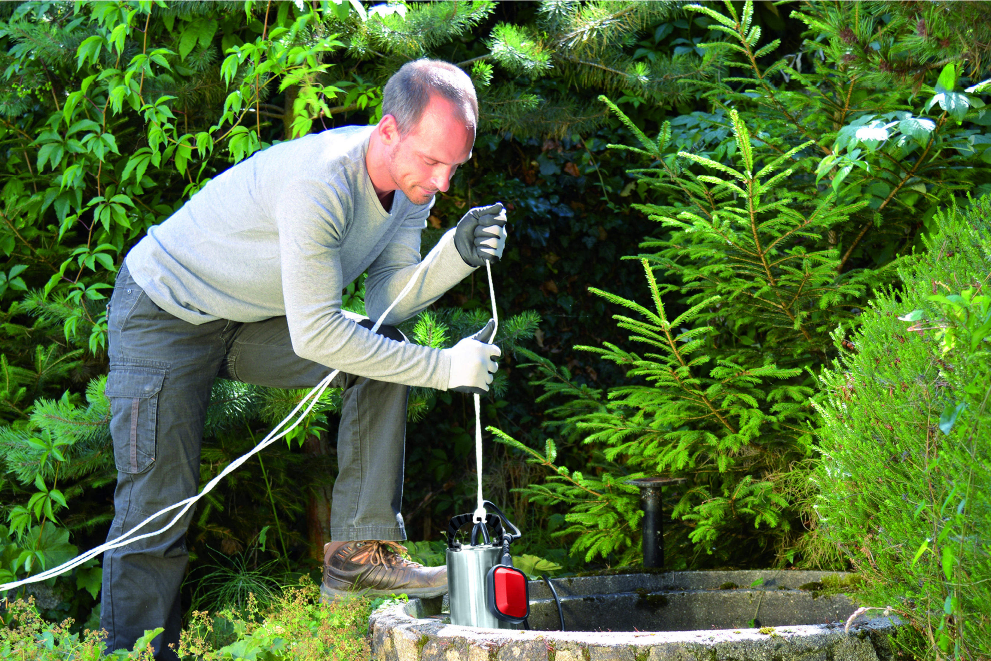 Mann im Garten arbeitet an einer Tauchwasserpumpe, in der Nähe eines steinernen Brunnens.