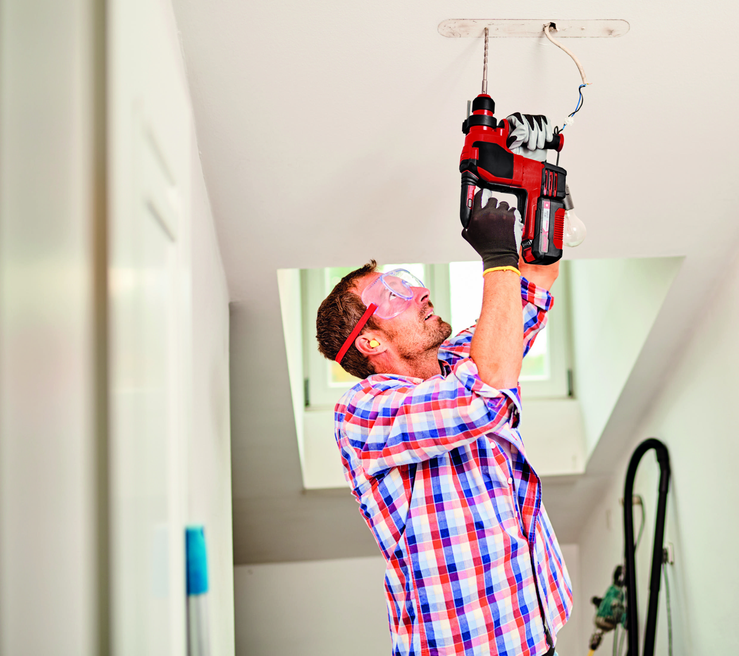Mężczyzna na drabinie instaluje lampę sufitową czerwoną wiertarką, nosząc okulary ochronne.